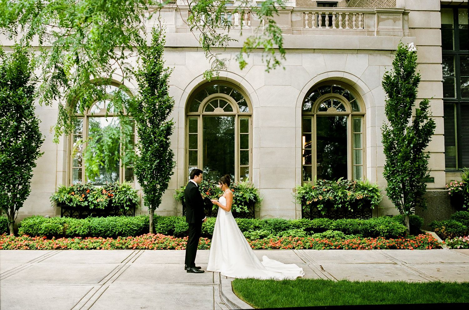 Bride and groom share a romantic first look outside an elegant stone building with arched windows and lush greenery.