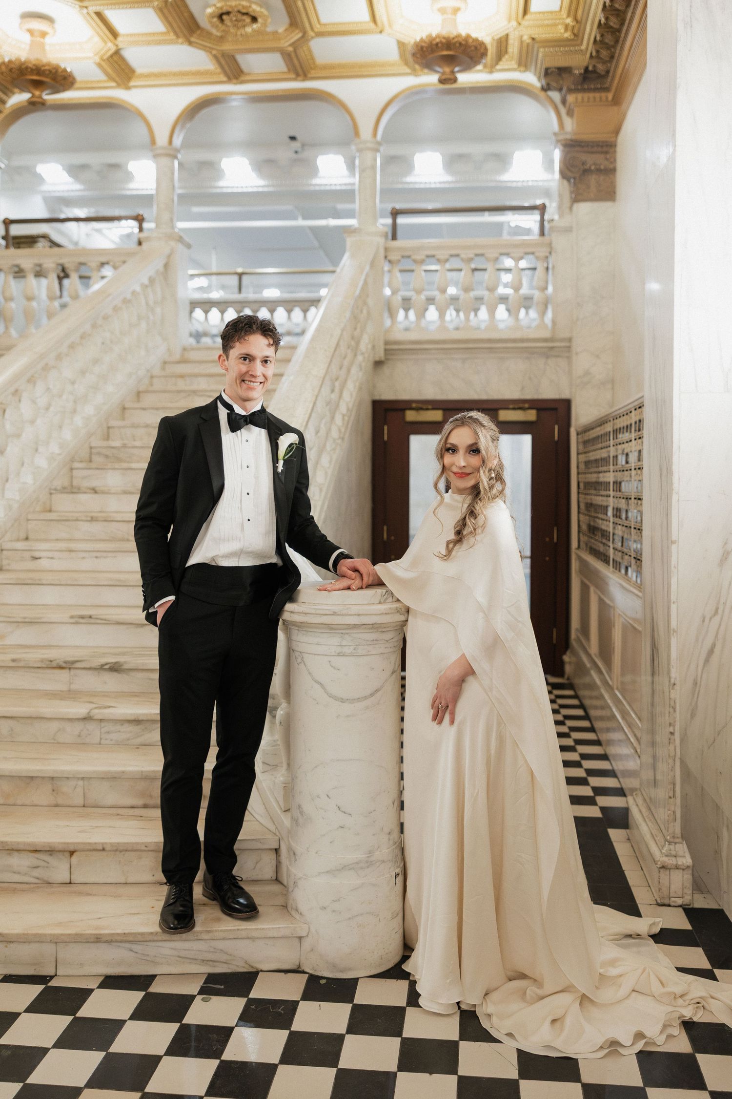 Newlyweds pose on a grand marble staircase with black and white checkered floors in an elegant venue.