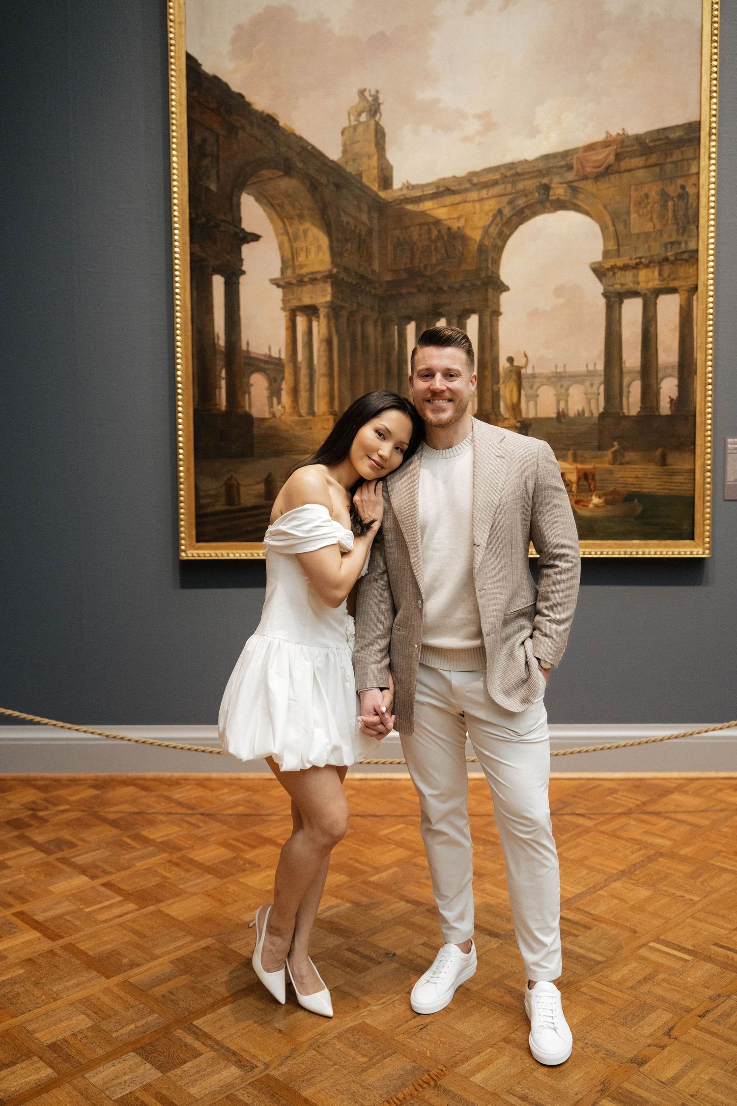Couple posing in front of a large classical ruins painting in an art museum, woman in white dress, man in beige suit.