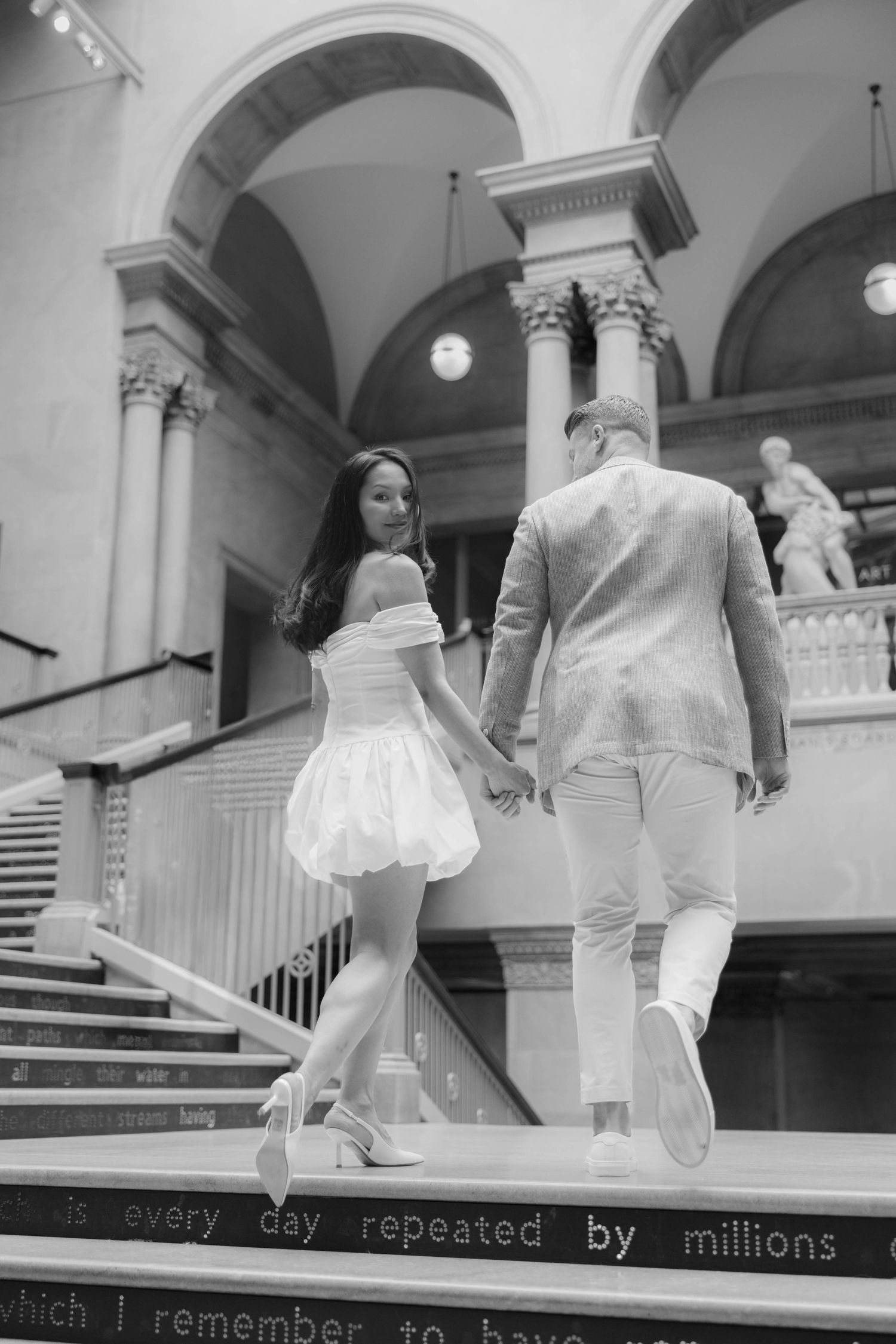 Couple holding hands walking up grand staircase in elegant museum, woman in white mini dress and heels.