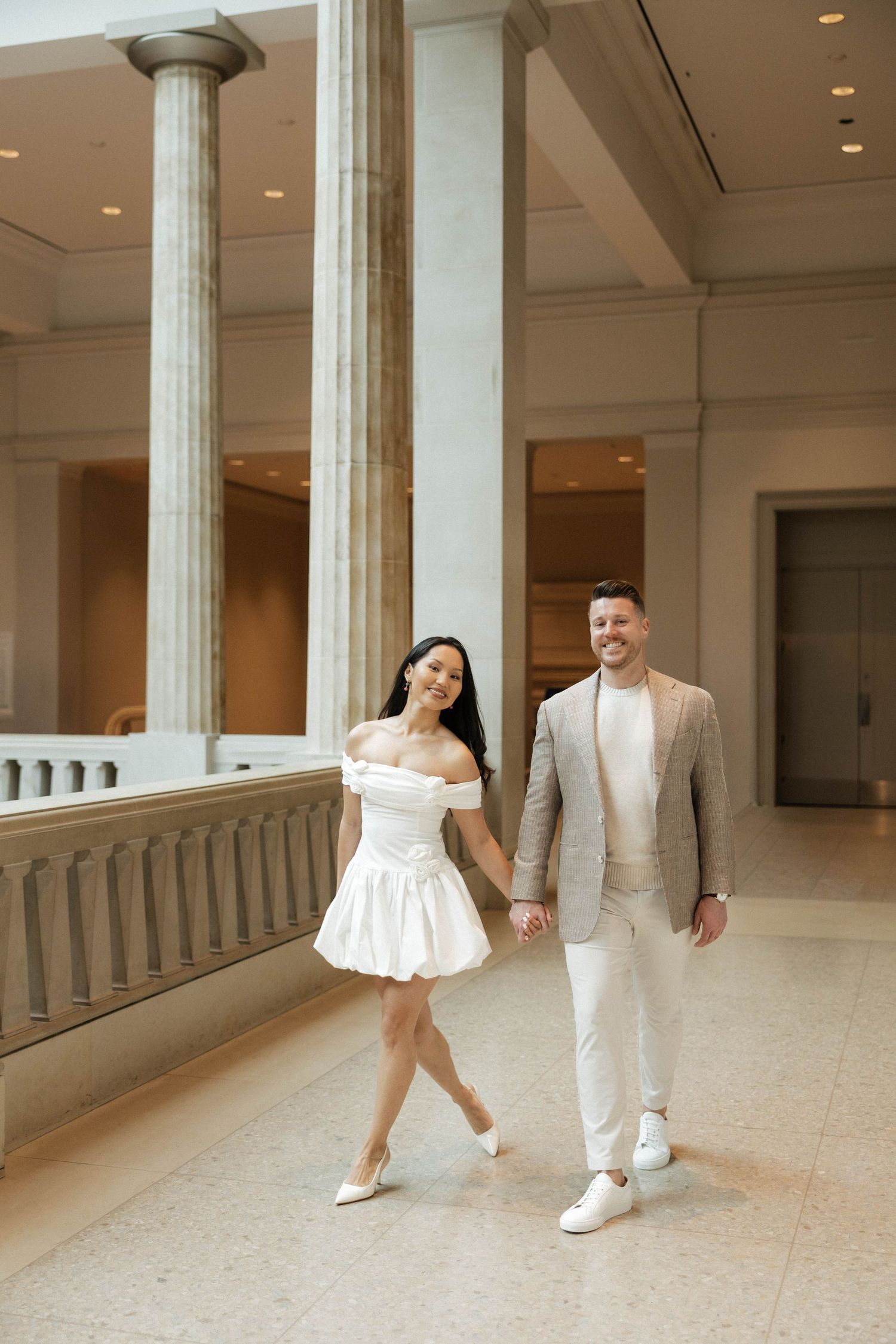 Stylish couple strolling hand in hand through a classical museum corridor with large marble pillars and marble flooring.