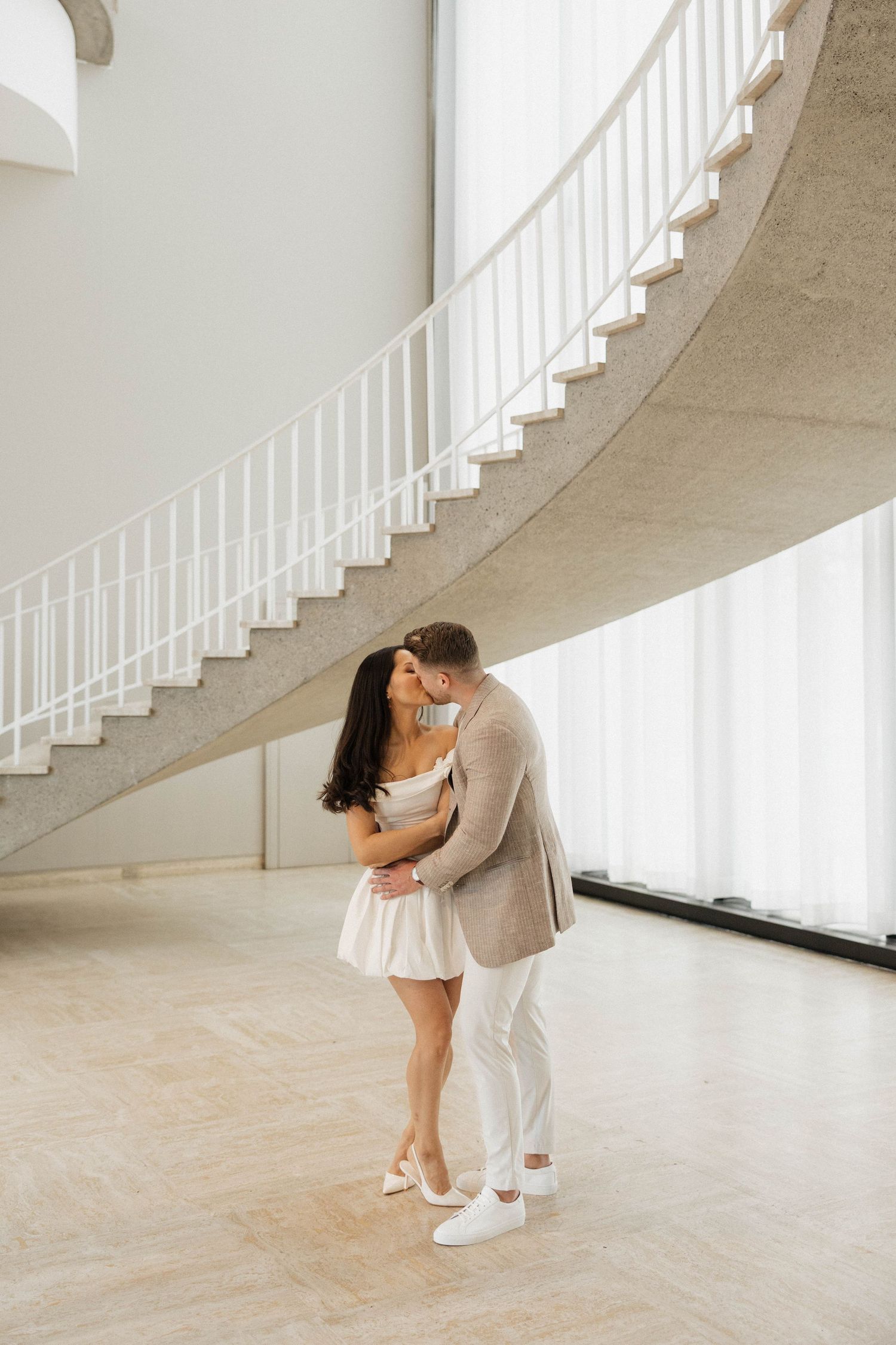 Couple sharing a romantic kiss beneath a curved white staircase in an elegant, bright interior space.