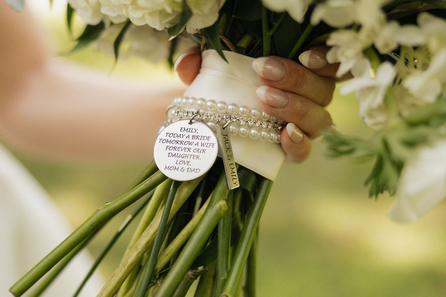 Bride holding white floral wedding bouquet with pearl bracelet and engraved memorial charm tag tied with ribbon.