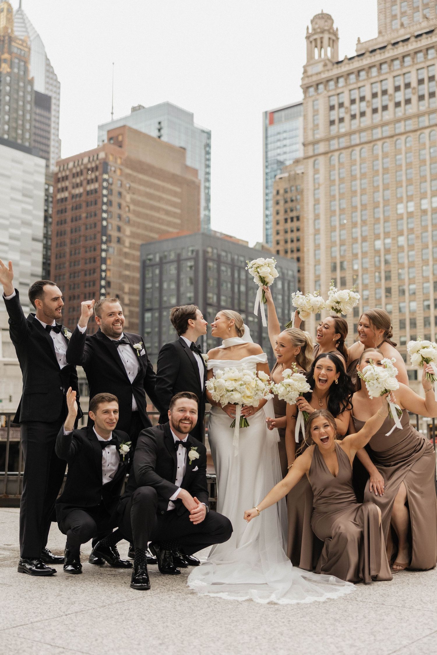 Joyful wedding party celebrating with raised bouquets against Chicago skyline backdrop.