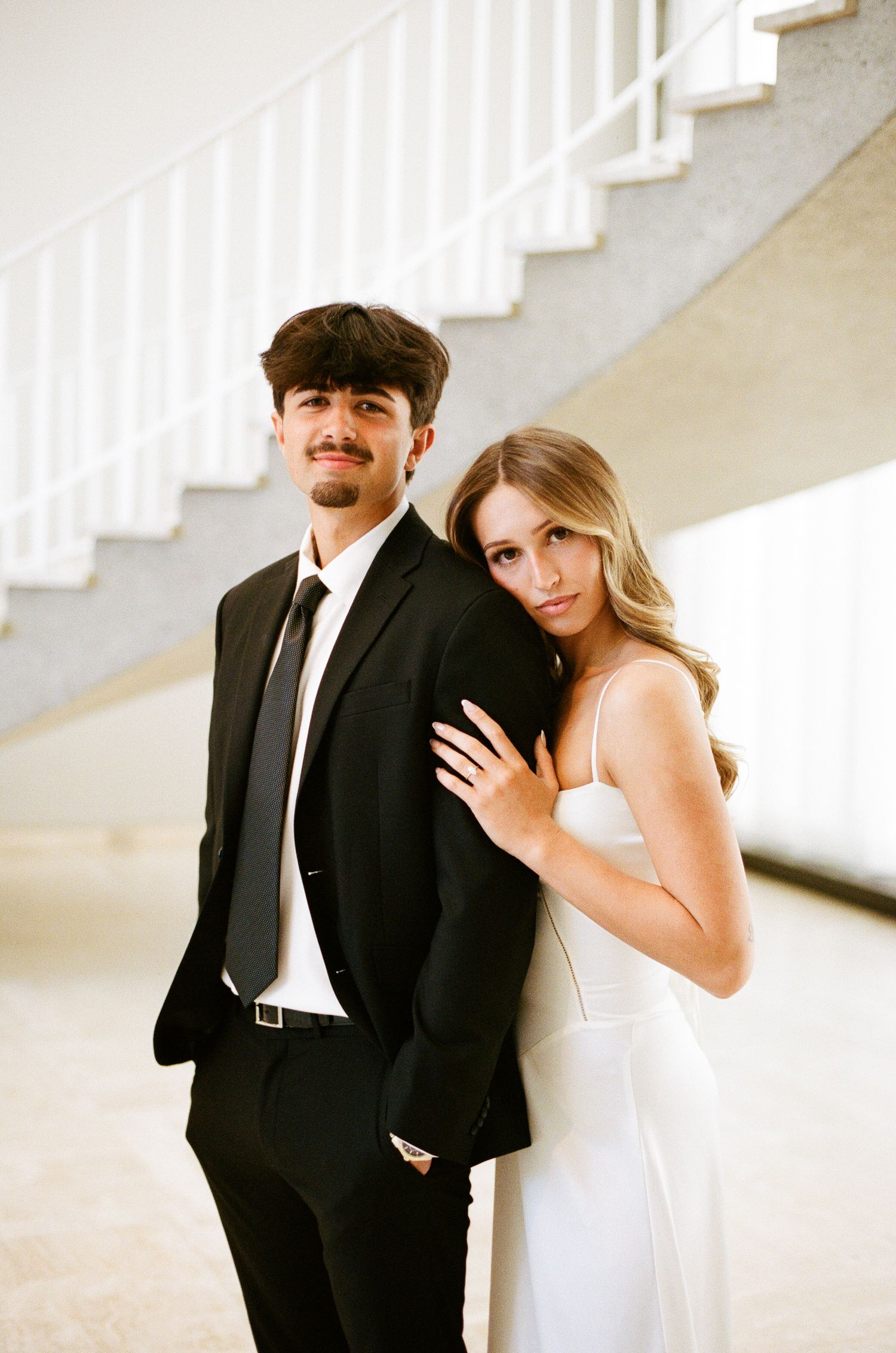 A young couple in formal attire pose elegantly near a spiral staircase, man in black suit, woman in white dress.