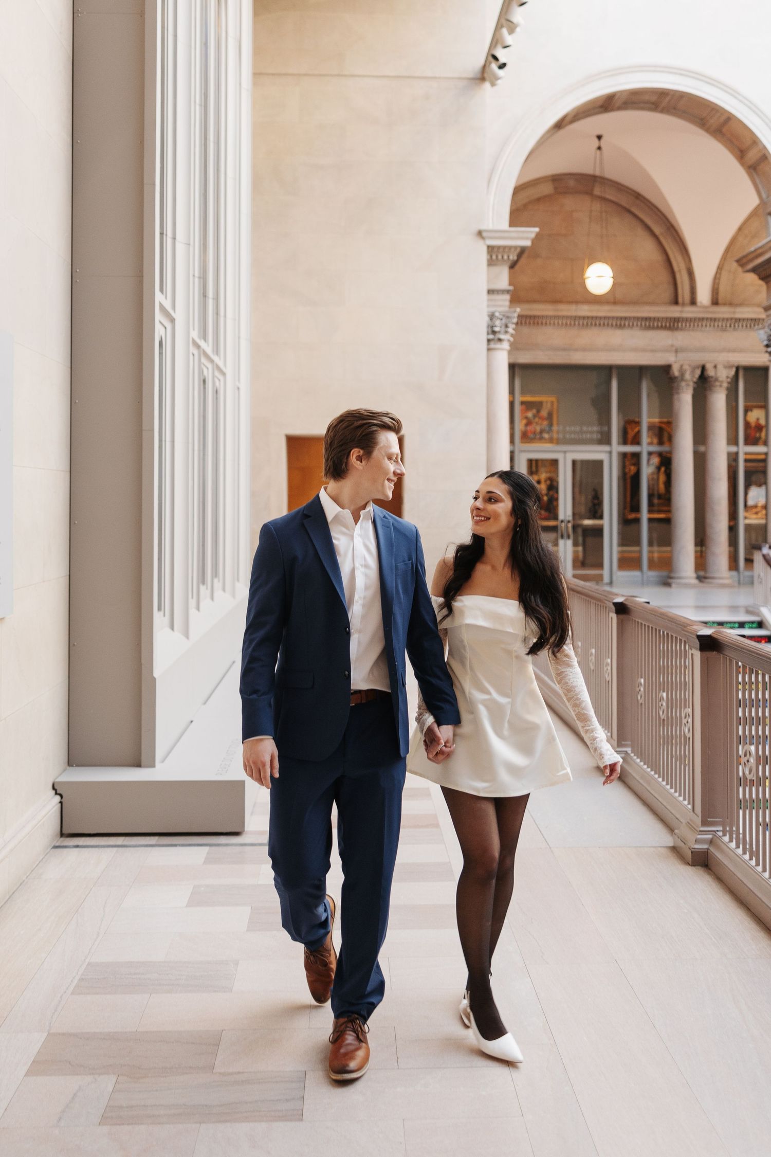 Couple walking together through an elegant colonnade with arched architecture.