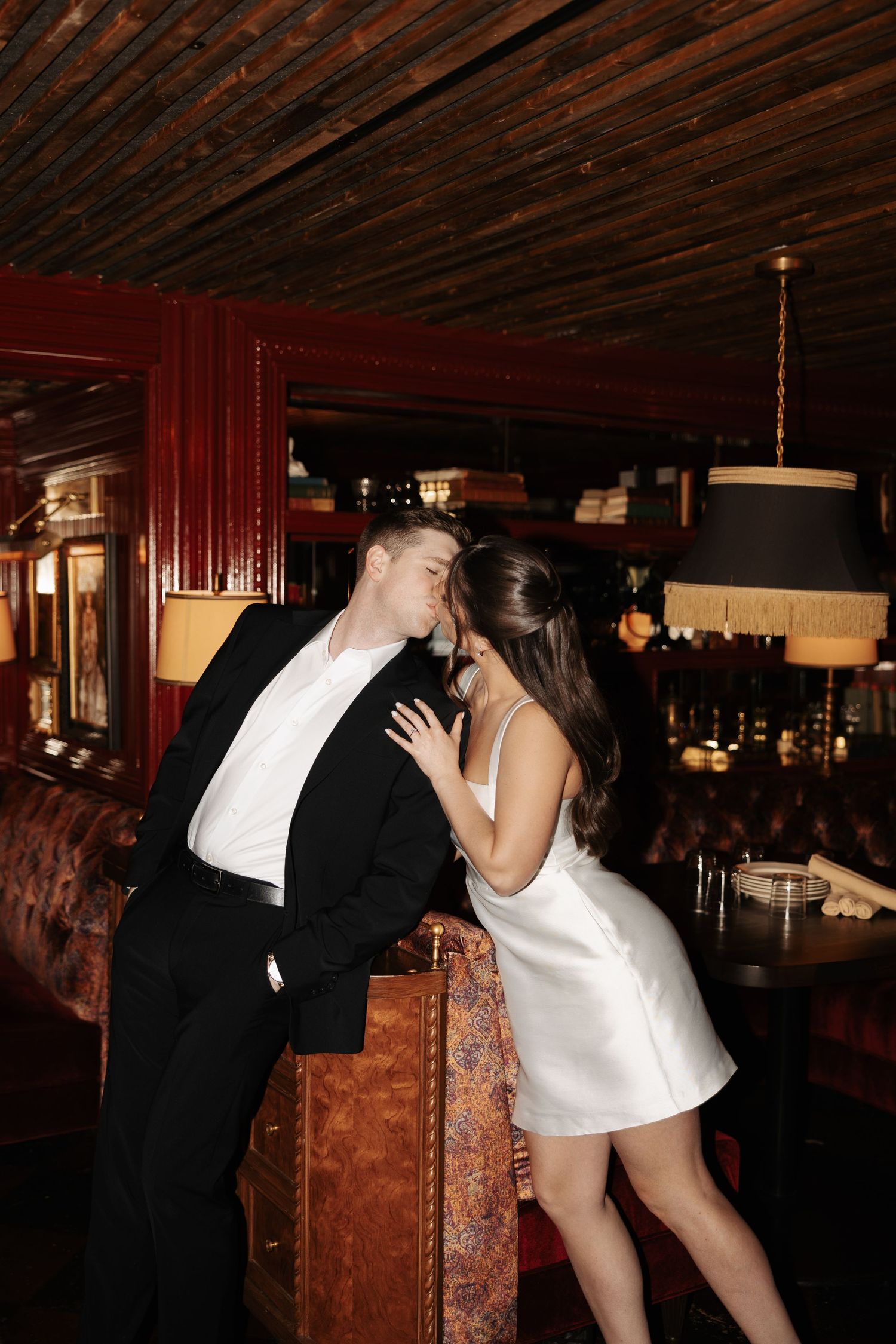A couple in formal attire share an intimate moment at a dimly lit bar with wooden decor and vintage ambiance.