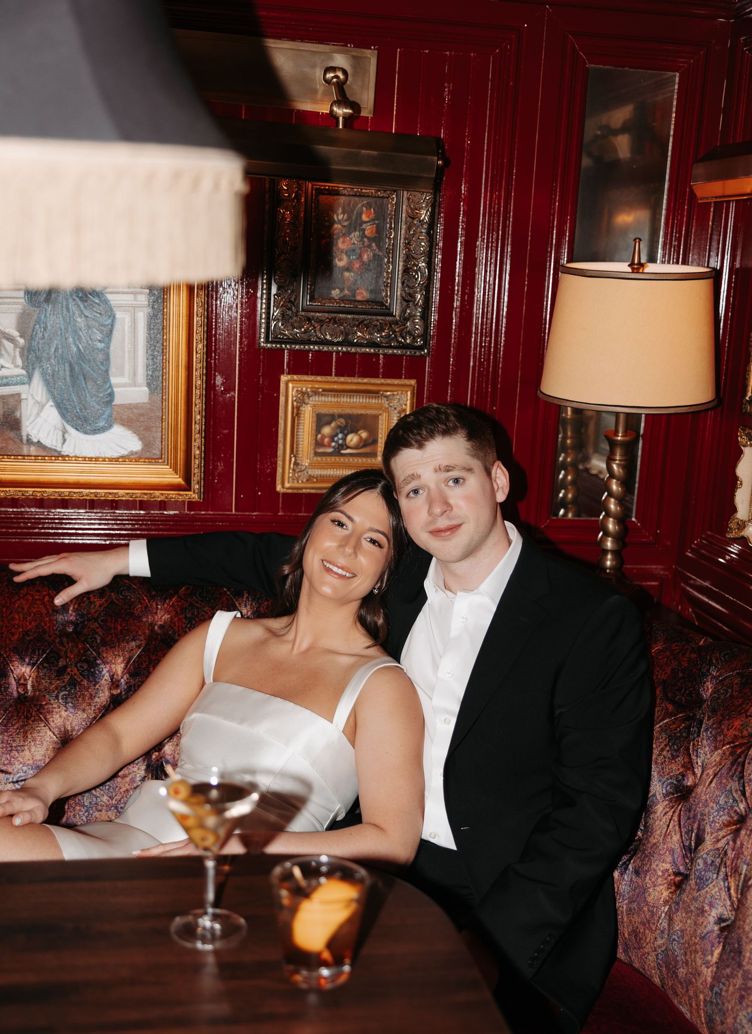 A couple in black and white formal wear sits together on a burgundy leather couch in a dimly lit bar.