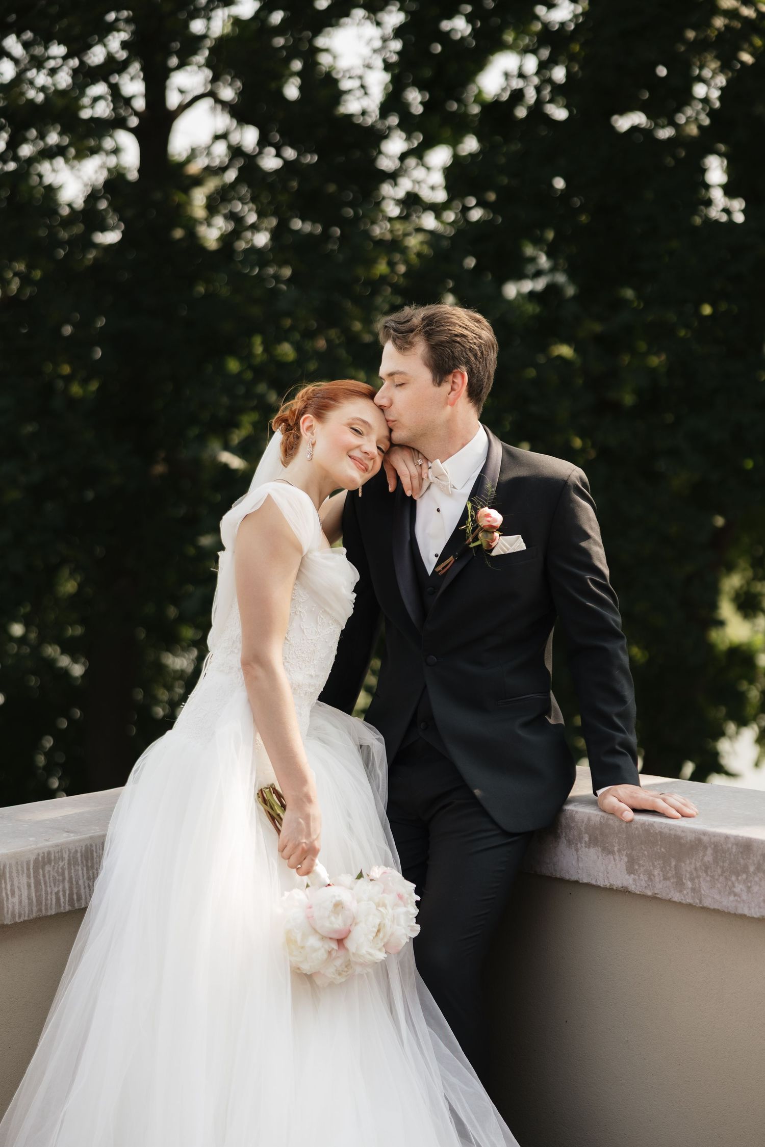 Groom in black suit kissing bride in white gown on cheek while she holds bouquet on outdoor terrace.