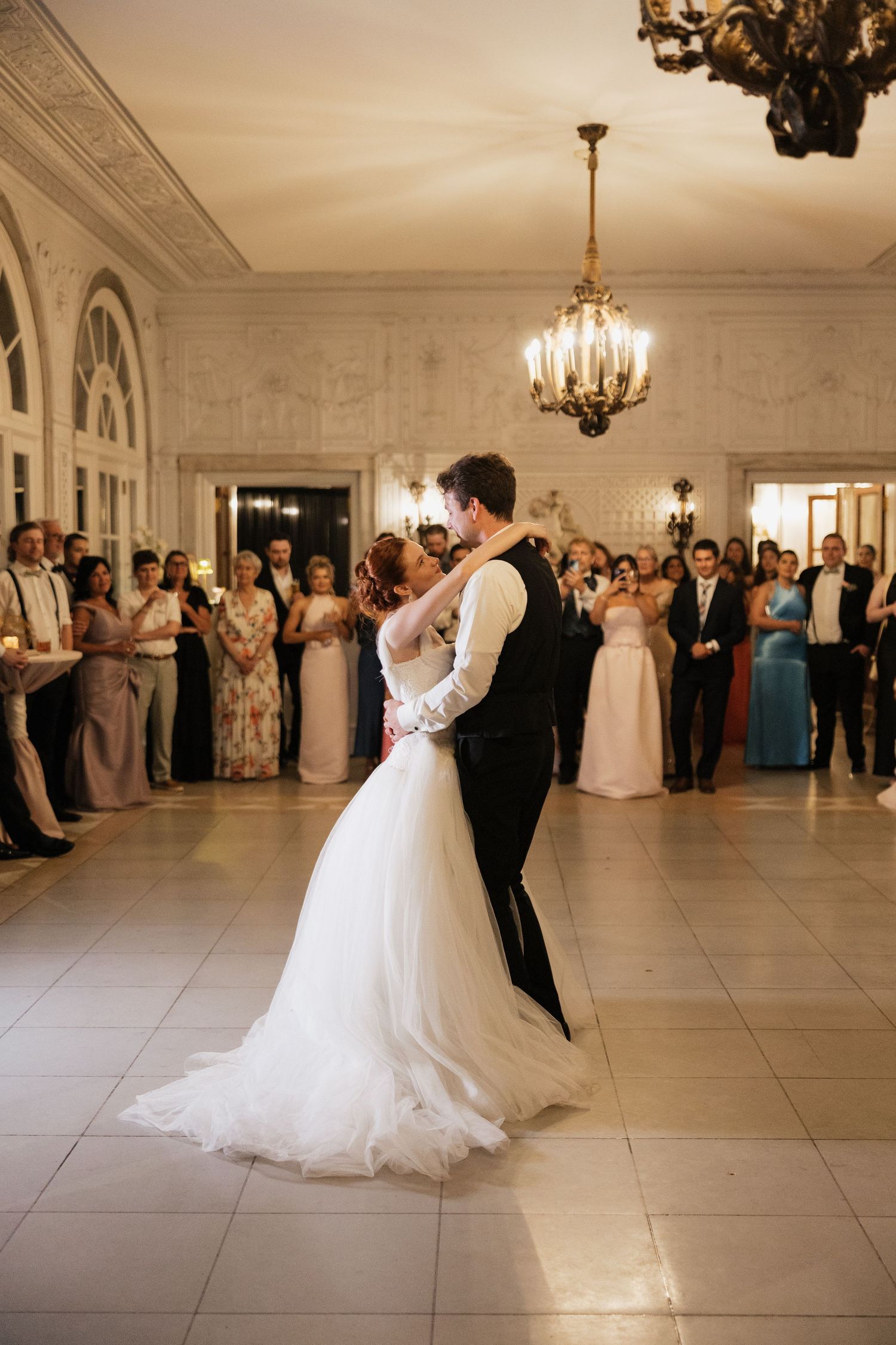 Bride and groom share their first dance in an elegant ballroom while wedding guests watch and celebrate around them.