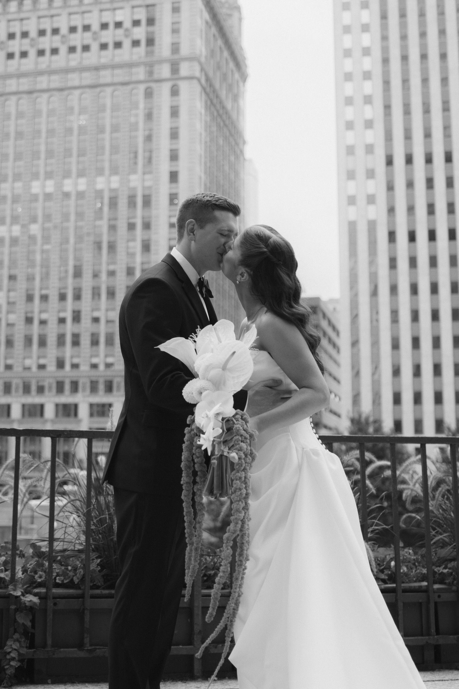Black and white photo of a bride and groom sharing a kiss on a city balcony, holding a cascading white bouquet before tall skyscrapers.