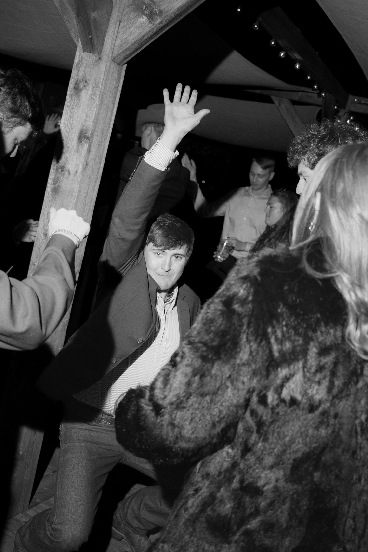Black and white photo of a smiling man in a blazer and open collar dancing with a raised hand at a lively party under wooden beams.