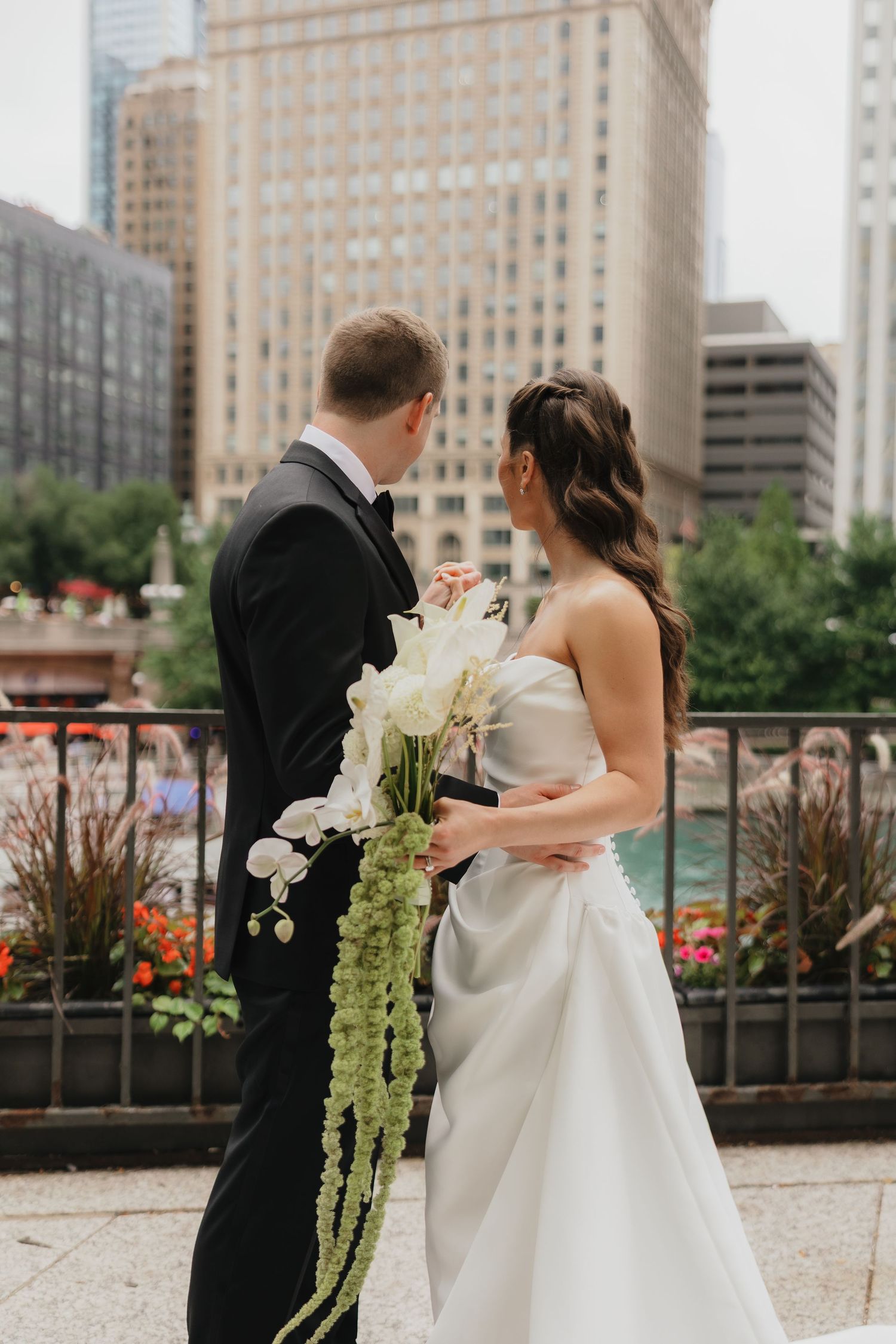 Bride in a strapless satin gown holding a cascading white calla lily bouquet beside a groom in a black tuxedo against a city skyline.