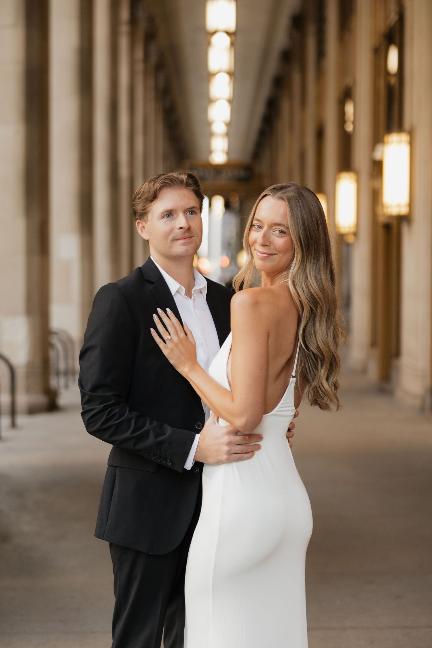 Couple in elegant attire embrace under a grand columned corridor with glowing lanterns, the woman showing her ring in an engagement photo.