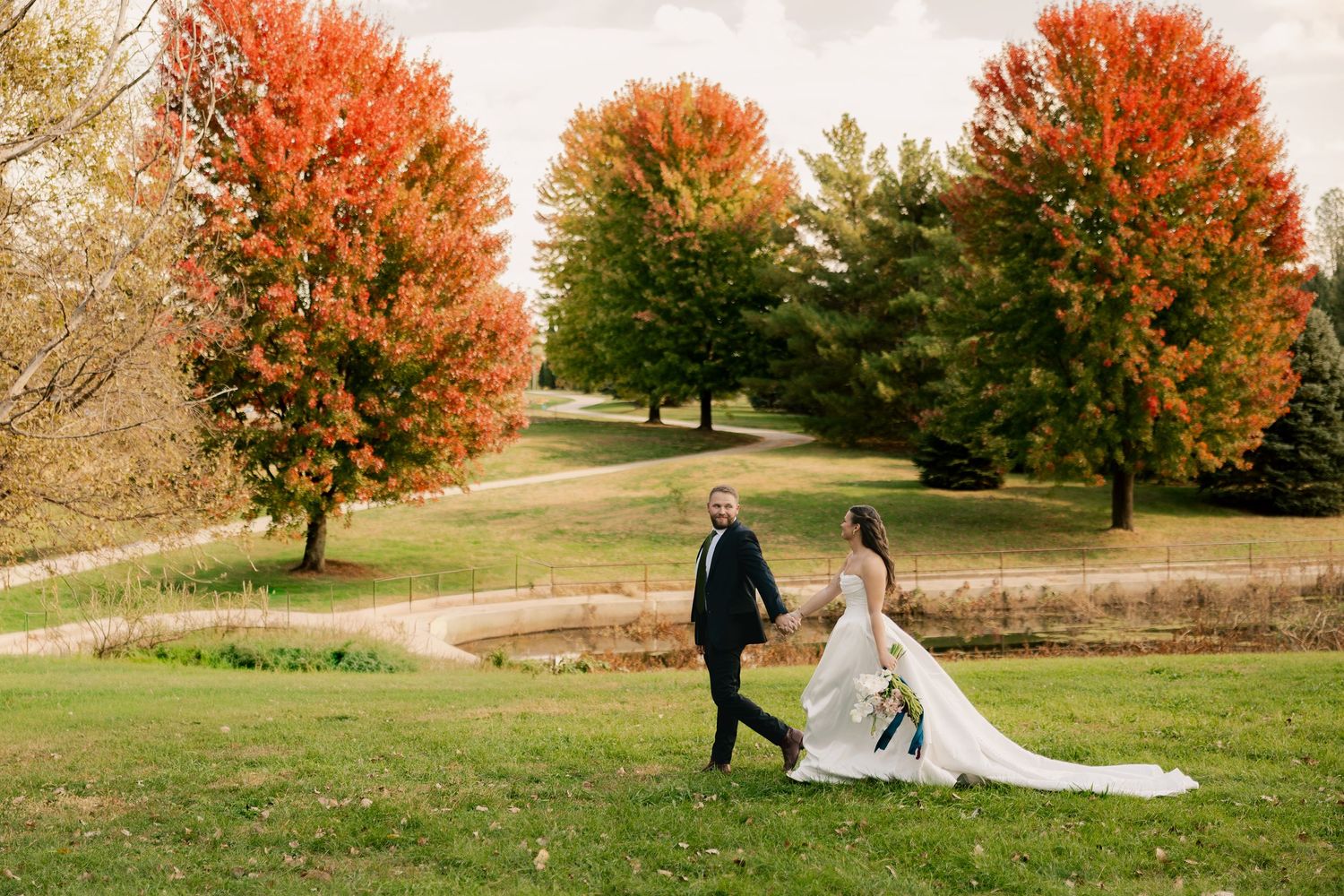 Bride in a long white gown and groom in a dark suit walk hand in hand across a grassy park lined with vibrant red and orange autumn trees.
