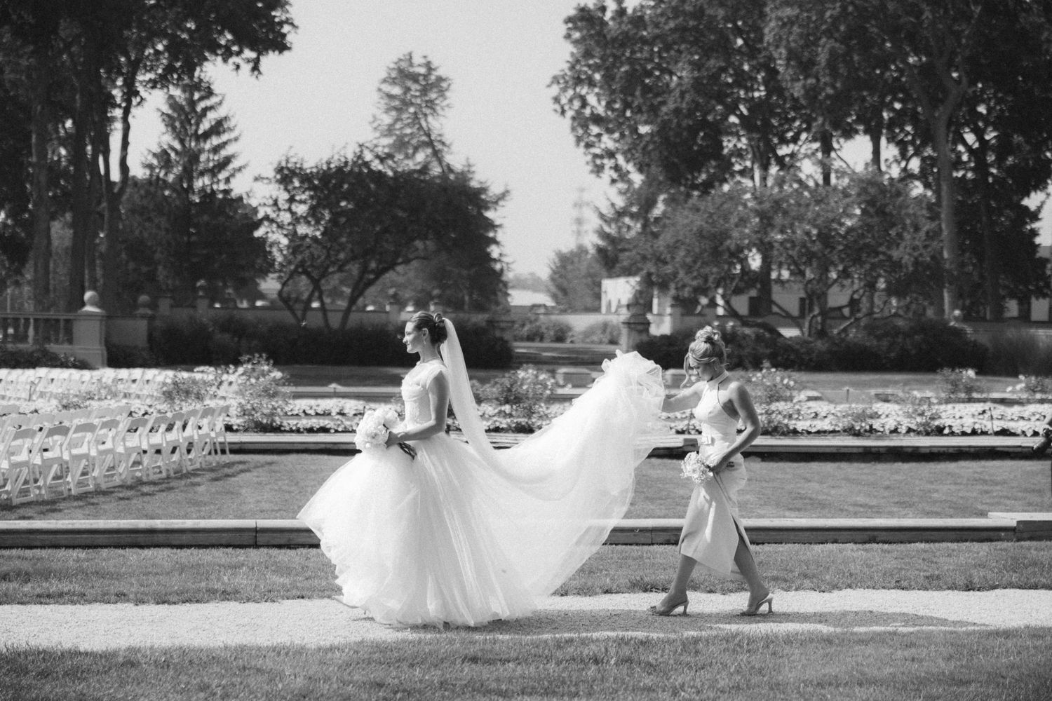 Black and white photo of bride in flowing gown with bridesmaid carrying train in formal garden.
