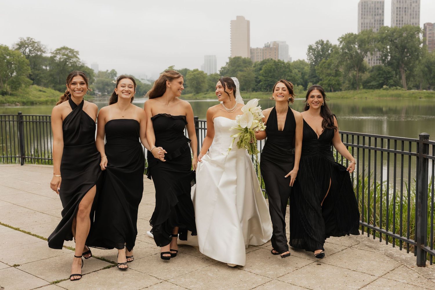 Bride in white gown walks with bridesmaids in black dresses near Central Park lake with NYC skyline backdrop.