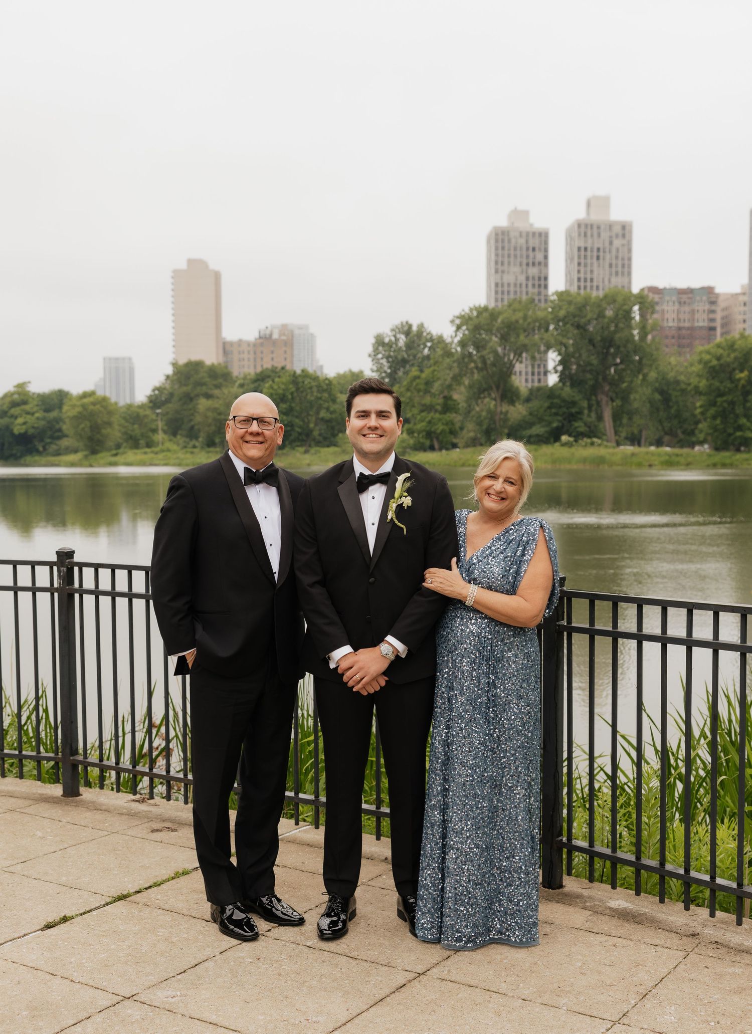 Groom in tuxedo poses with parents by a lakeside railing, city skyline and lush greenery in the background.