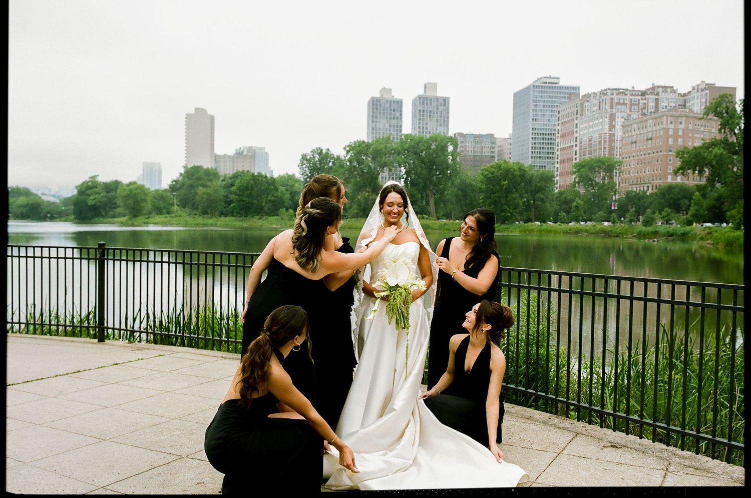Bride in white gown with bridesmaids in black dresses by a lake in Central Park, NYC skyline in background.