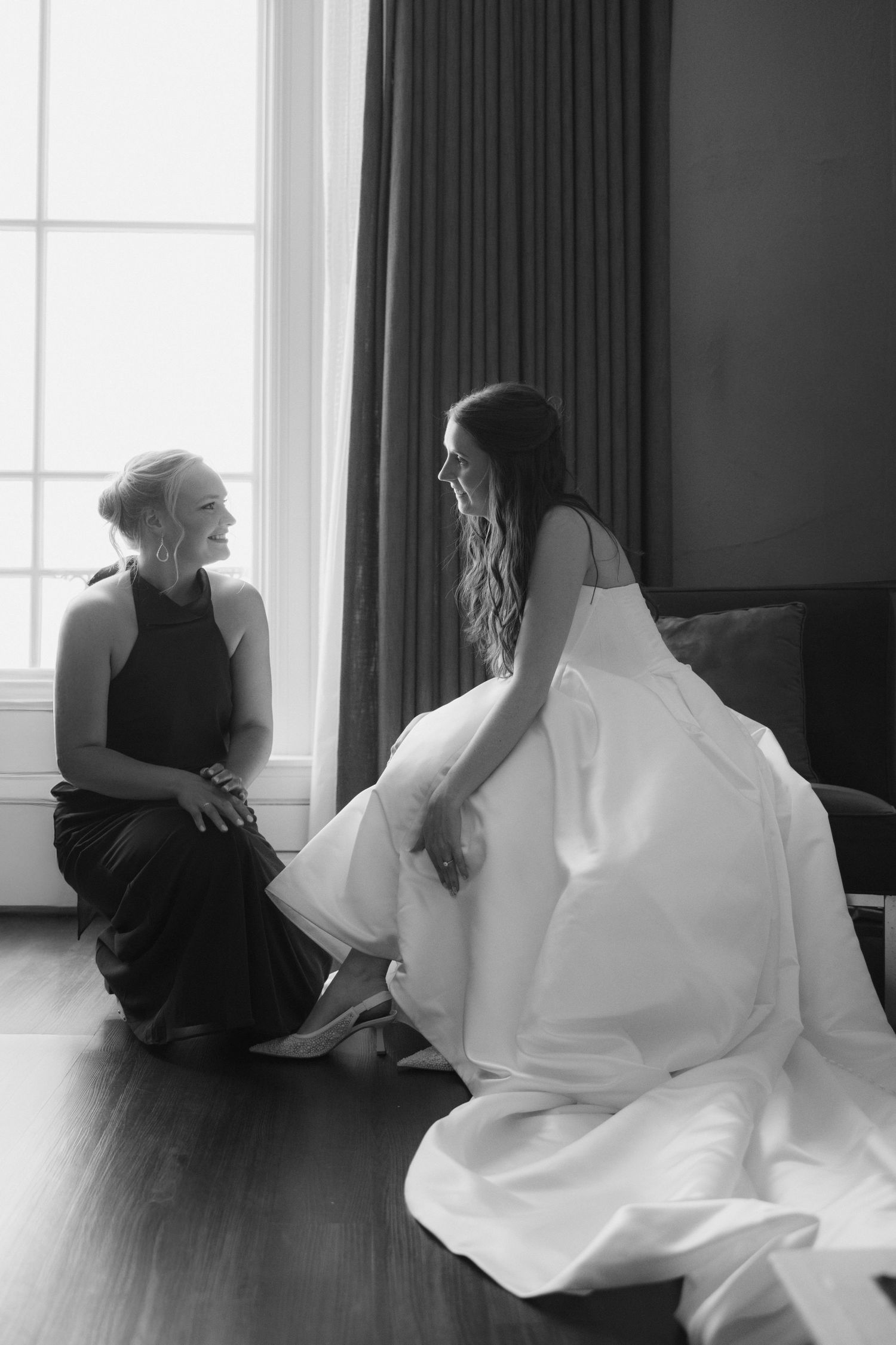 Black and white photo of a bride in white gown and bridesmaid in black dress sitting on floor chatting by a window.