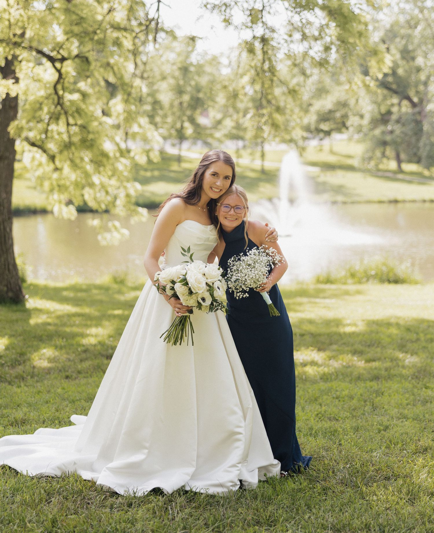 Bride in white strapless gown holds bouquet beside woman in navy dress near fountain in lush green park.