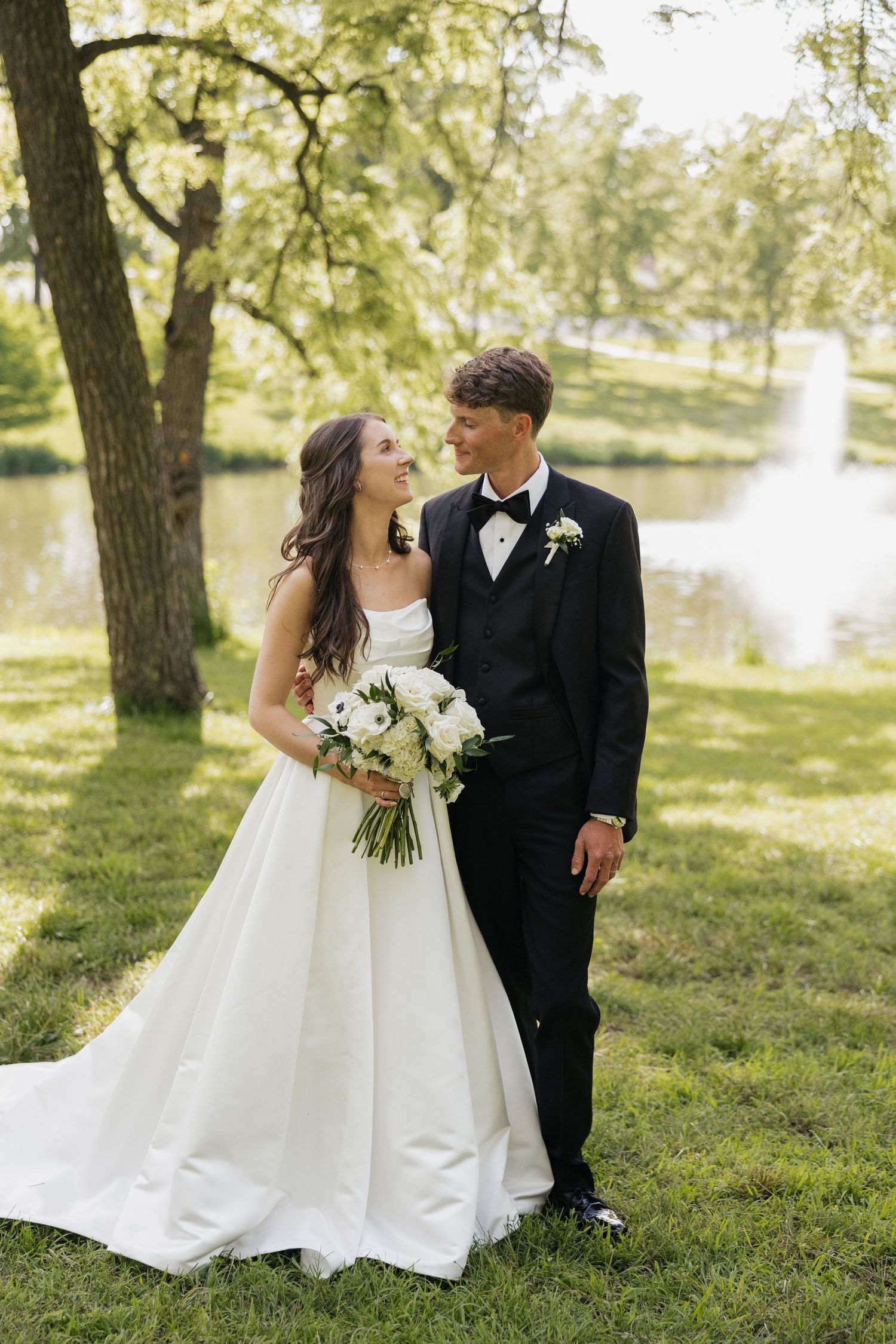 A bride and groom share a romantic moment outdoors near a pond, she holds white flowers in a ball gown, he wears a black tuxedo.