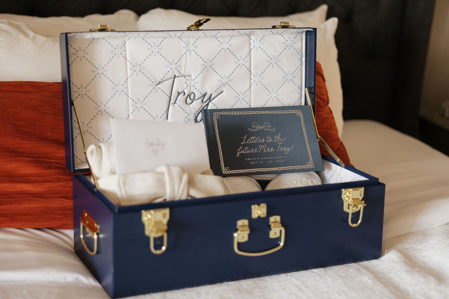 Navy blue keepsake trunk gift box personalized with Troy name, containing letters and white items on hotel bed.