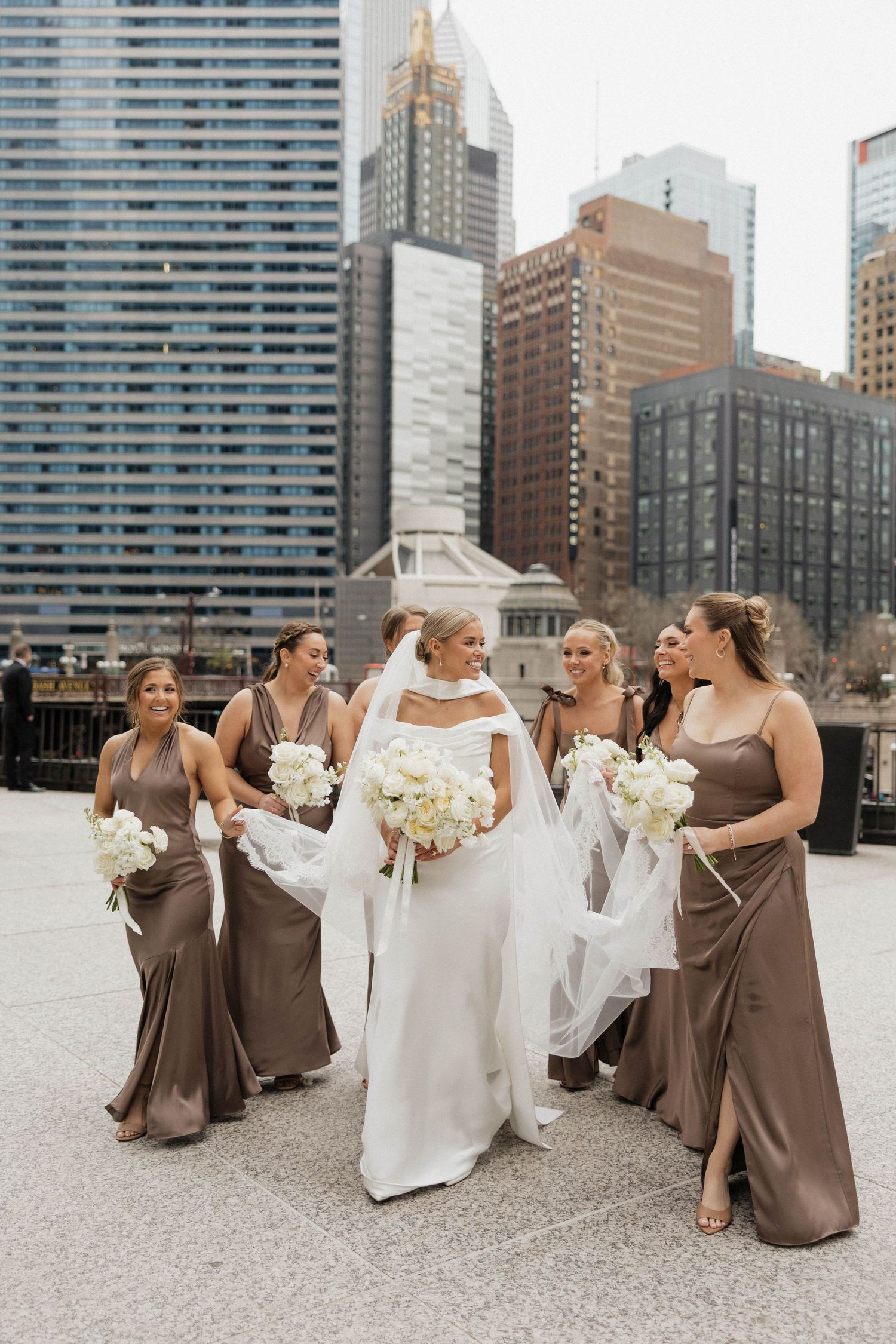 Bride in white gown with bridesmaids in taupe dresses holding white bouquets with city skyline.