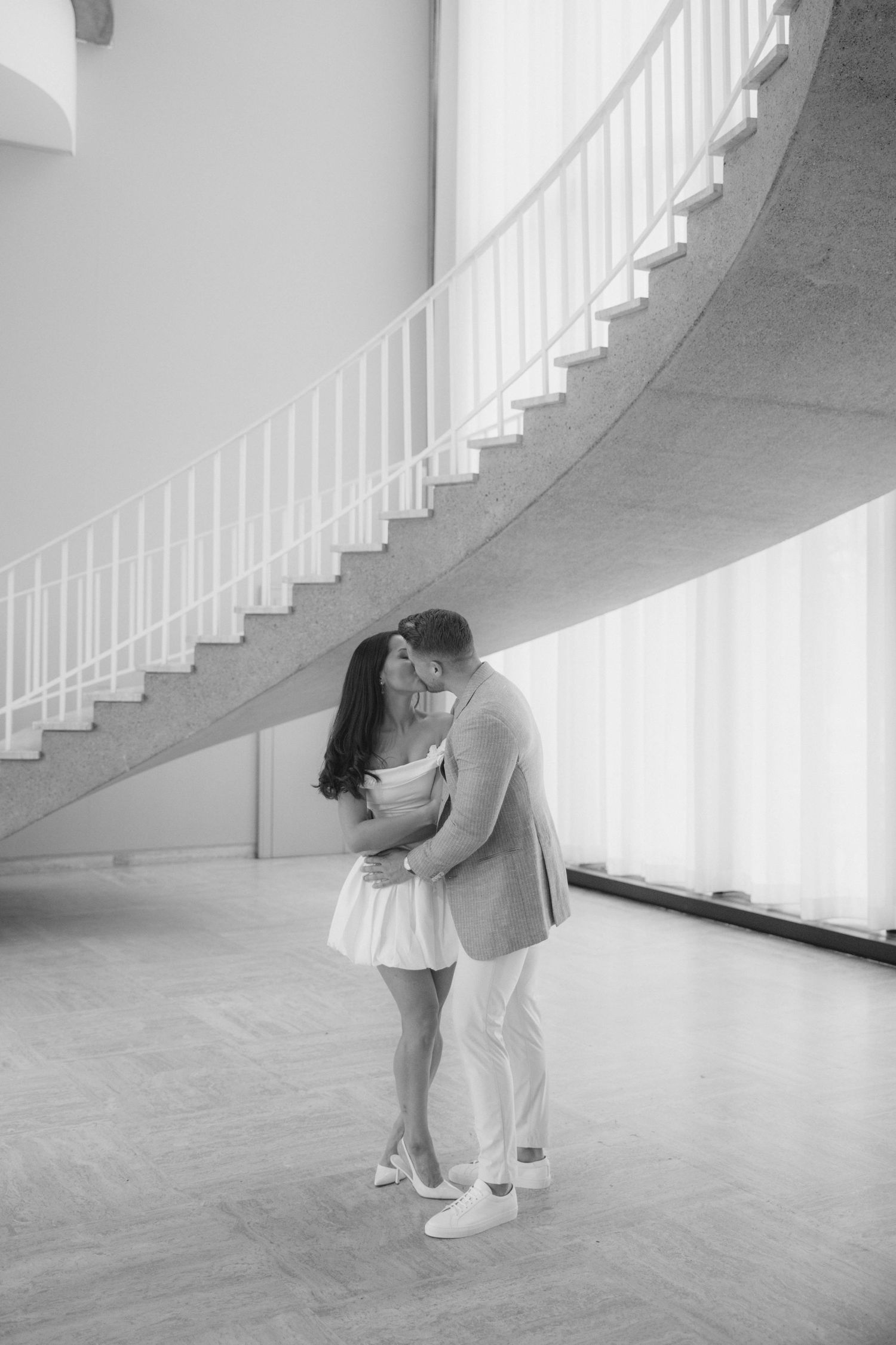 Black and white photo of a couple kissing under a curved staircase in an elegant indoor space.