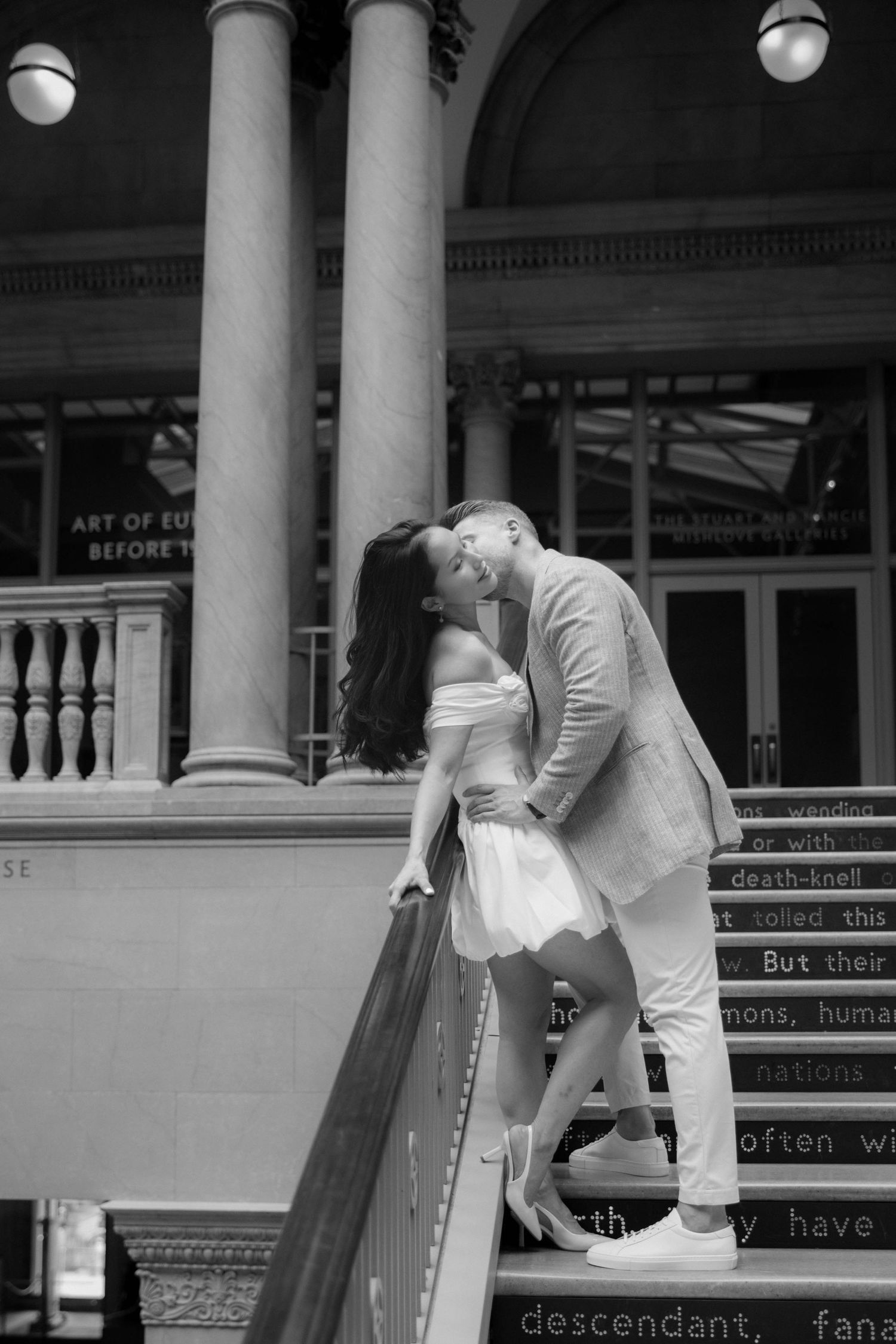 Black and white photo of a couple sharing a romantic kiss on grand staircase steps with tall columns behind them.