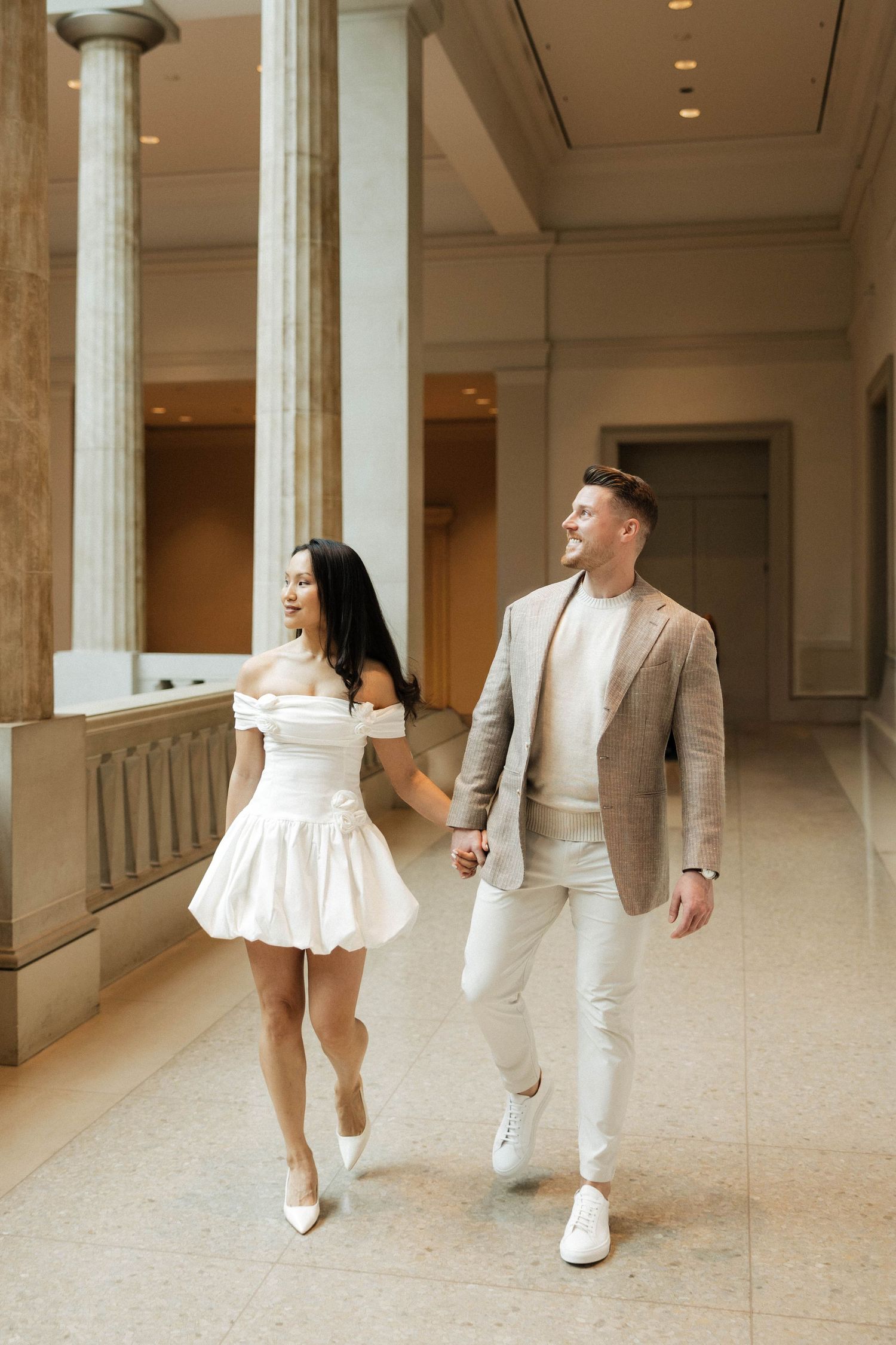 Fashionable couple in coordinated neutral outfits walking hand in hand through a classical architecture museum hallway.
