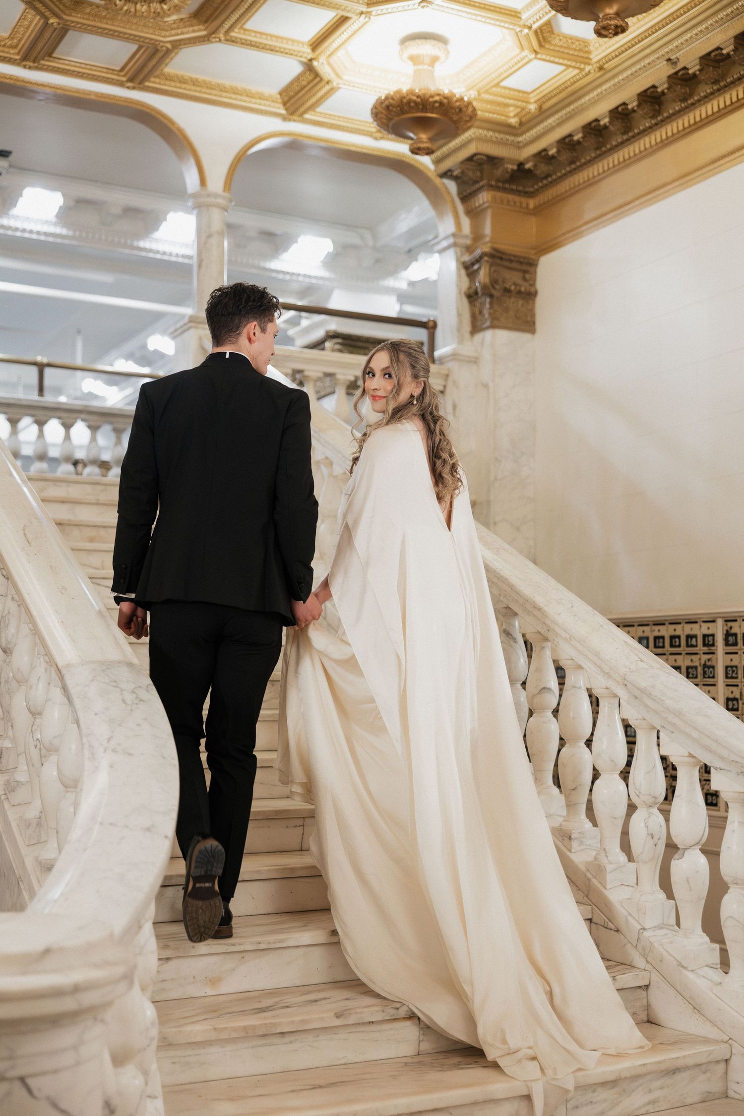 Newlyweds descending ornate marble staircase with flowing white dress trailing behind.