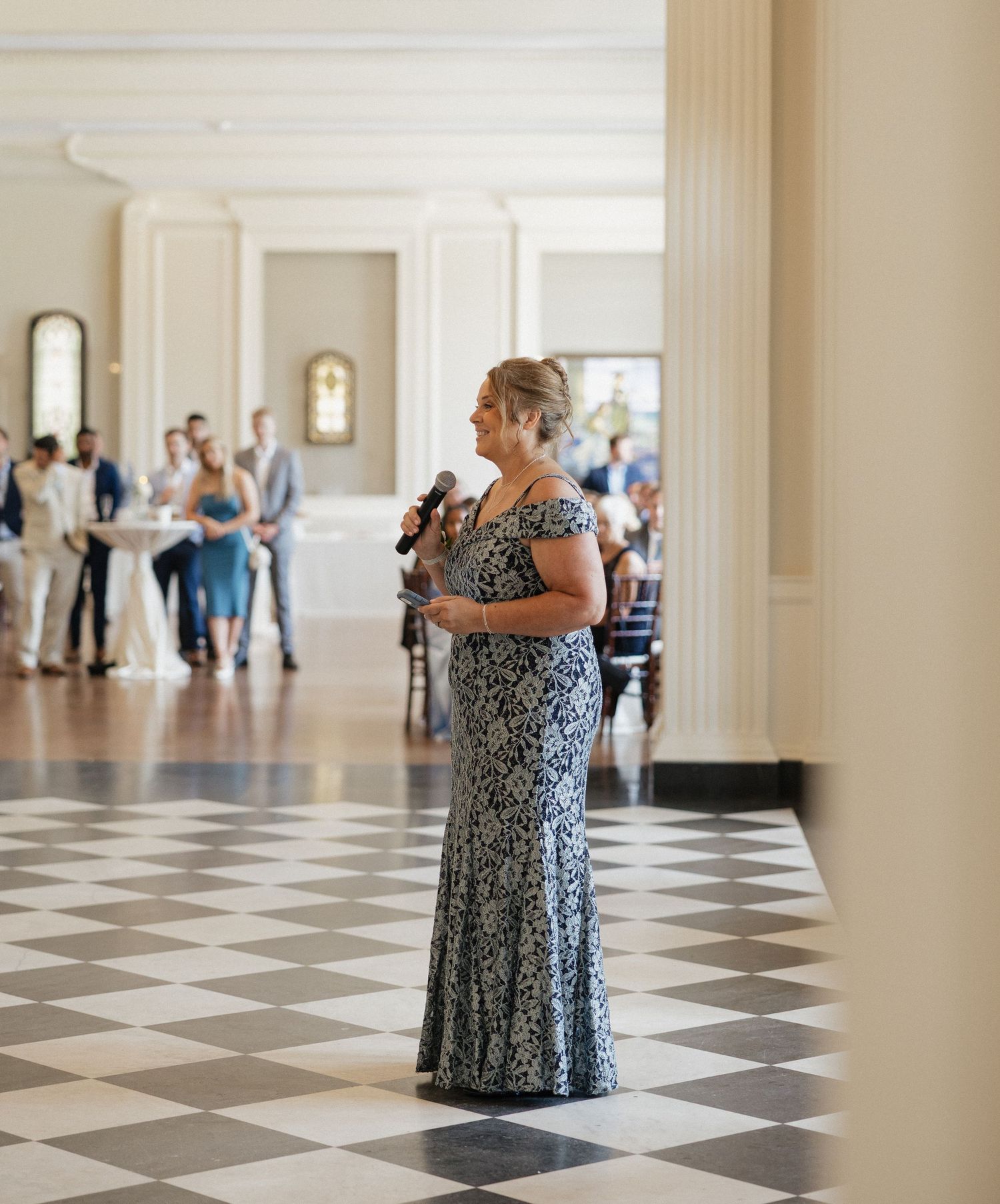 A person in a floral dress stands in a large ballroom with black and white checkered floors and tall columns.
