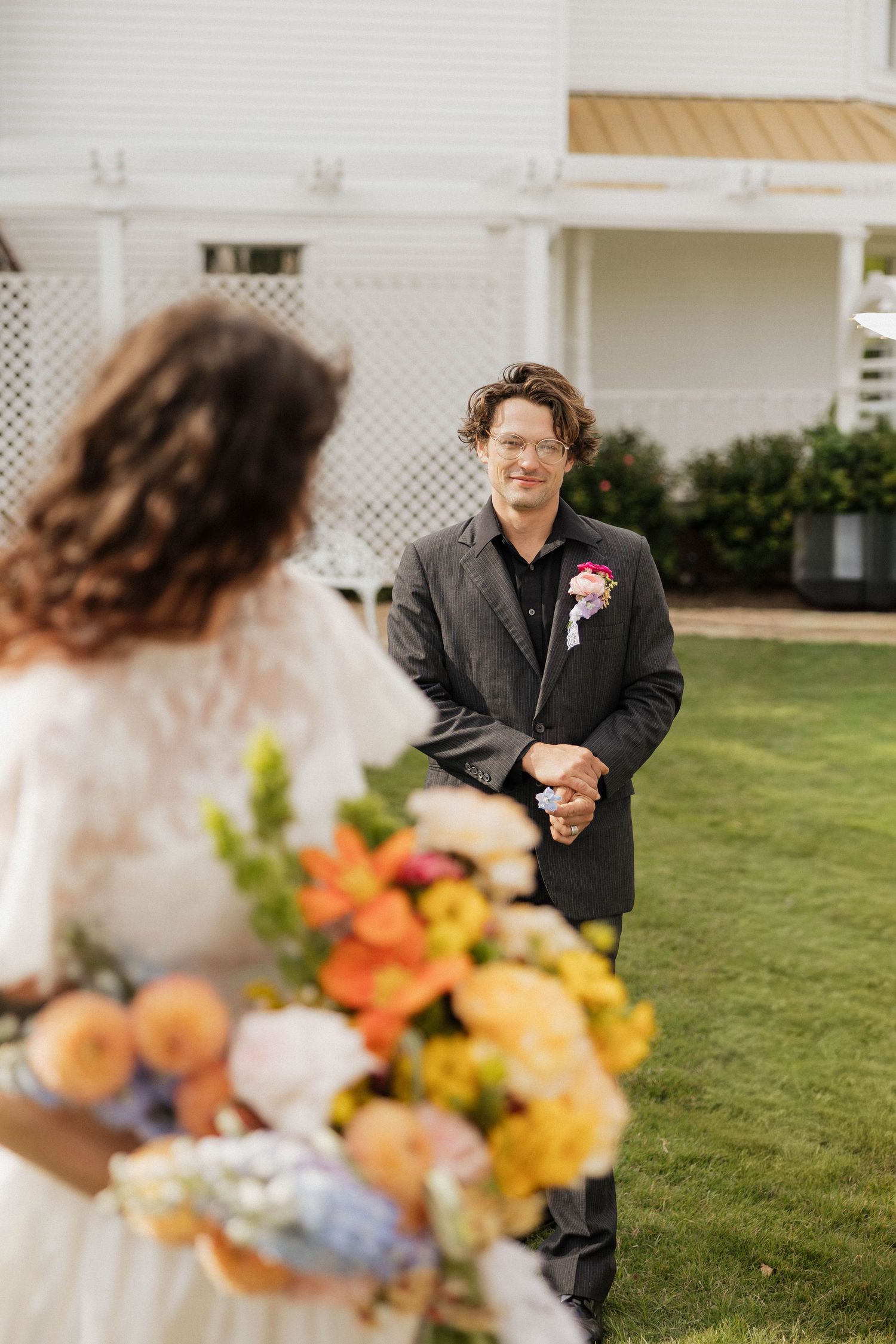 Groom in black suit smiling at bride holding colorful bouquet during outdoor wedding ceremony.