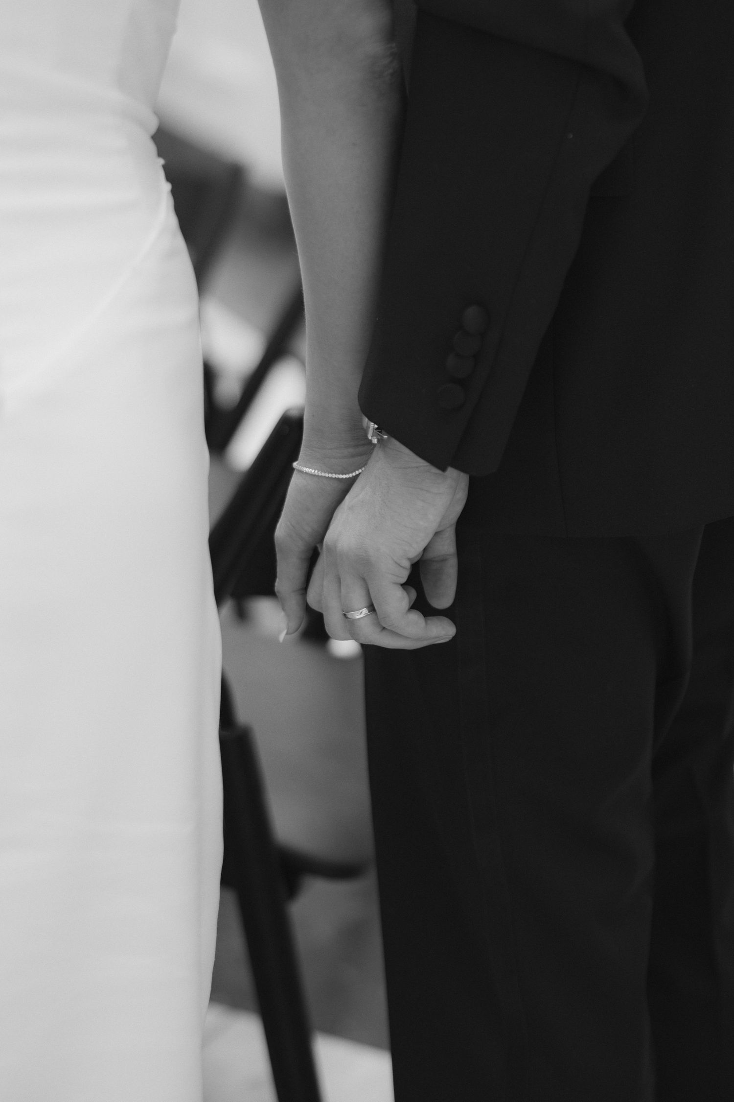 Black and white photo of bride and groom holding hands at wedding ceremony, showing rings and elegant attire.