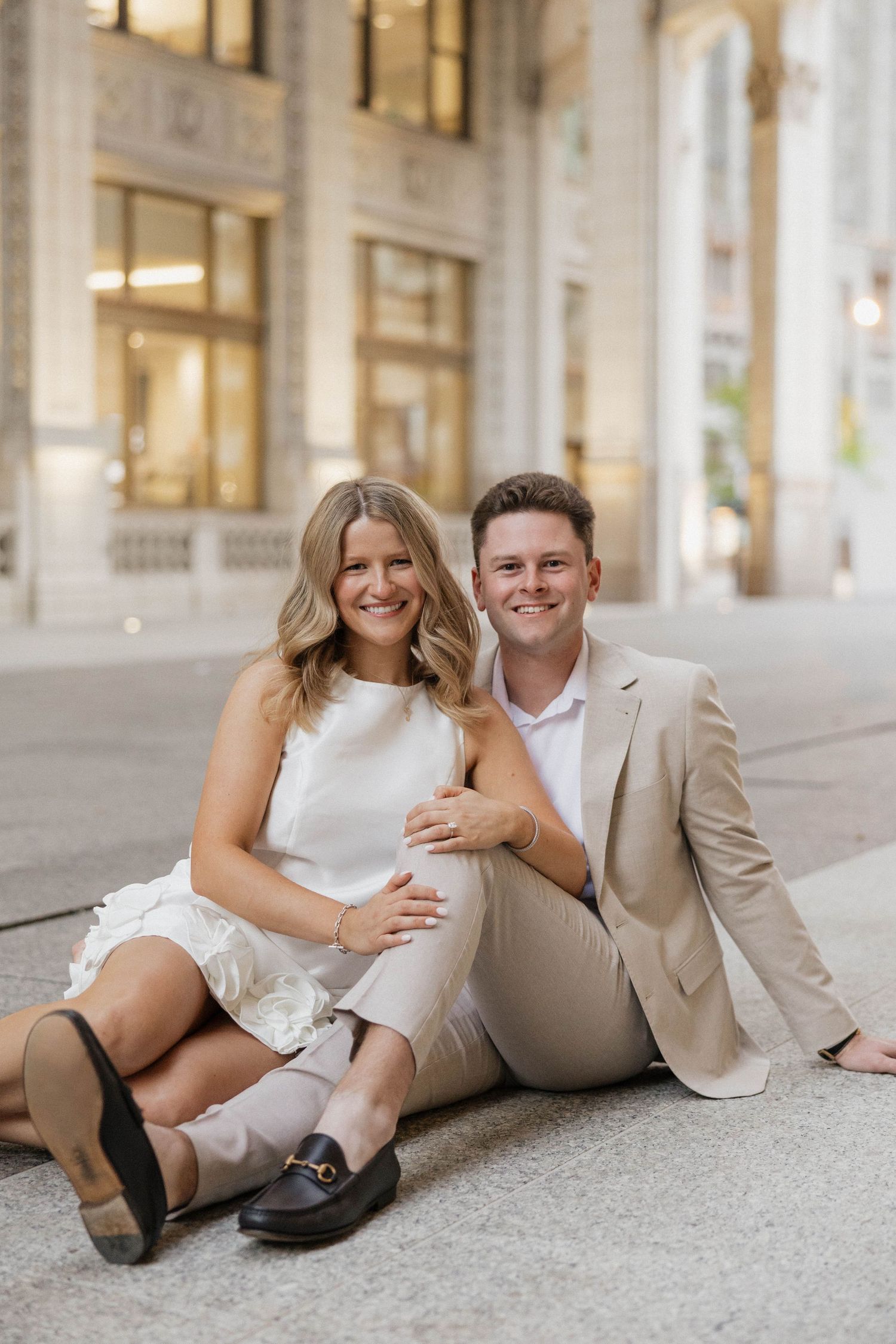 Engaged couple sitting on urban steps, woman in white dress and man in beige suit, smiling warmly.