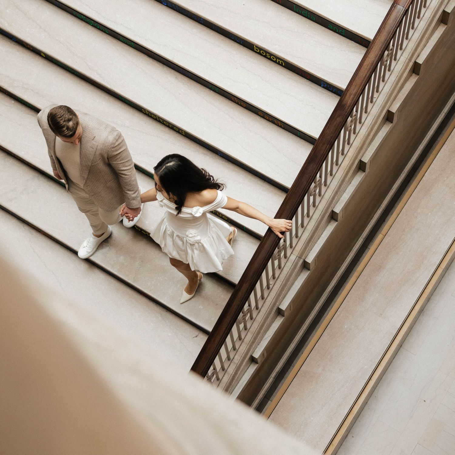 Aerial view of a couple holding hands, man in beige suit and woman in white dress, walking up stairs.