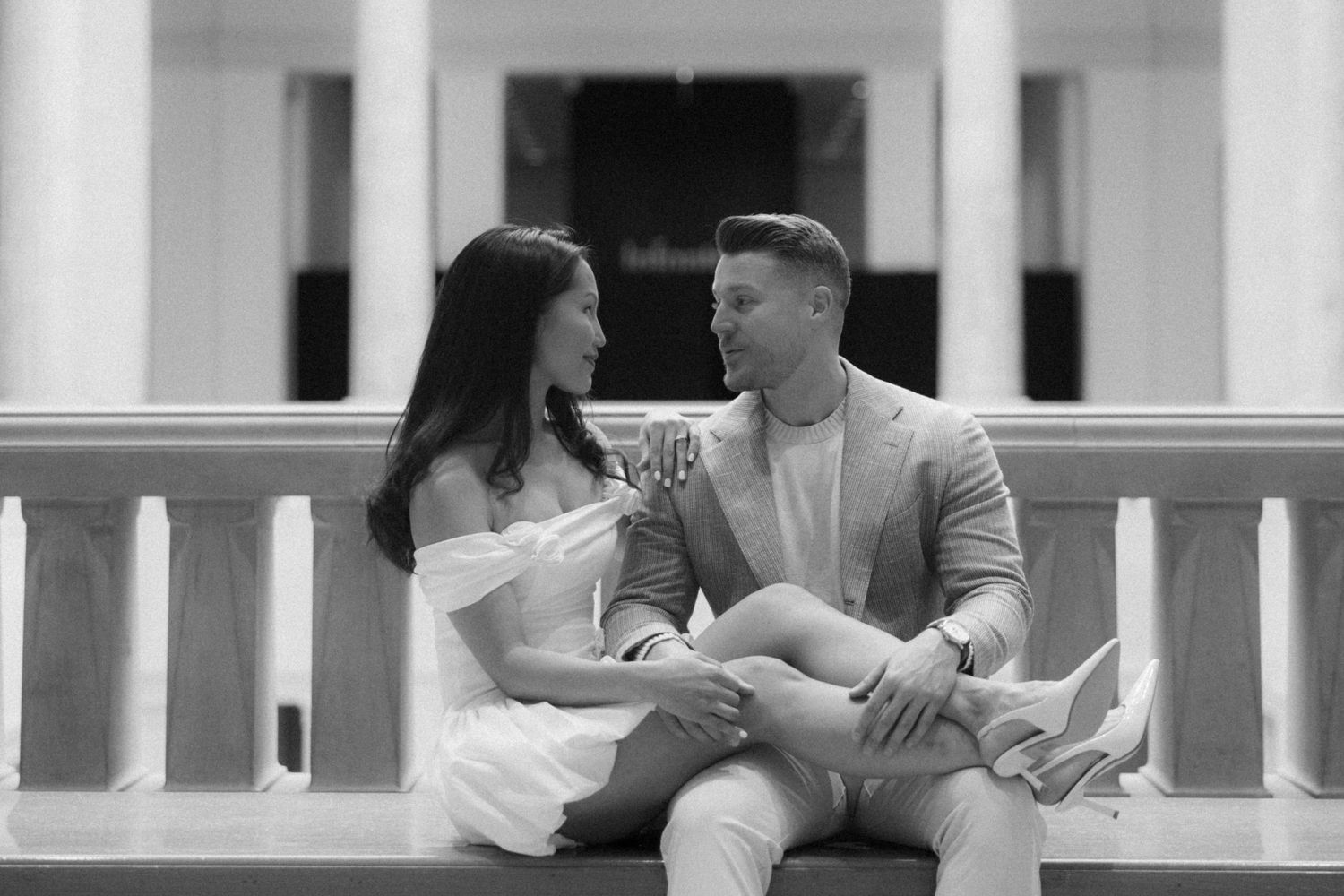 Black and white photo of a couple sitting on a bench, gazing at each other during an engagement session.
