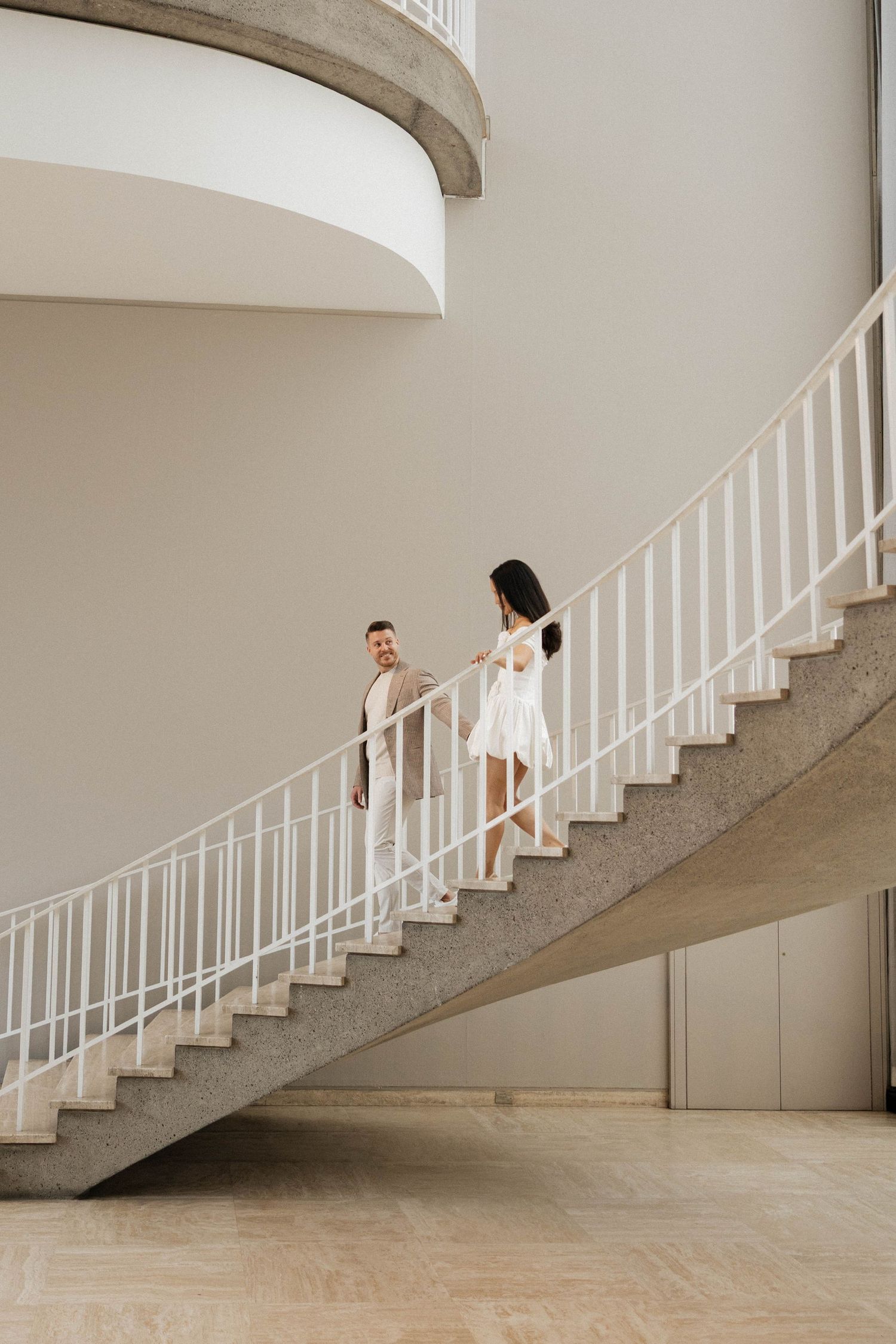 Couple holding hands on sweeping curved staircase in modern beige interior space.