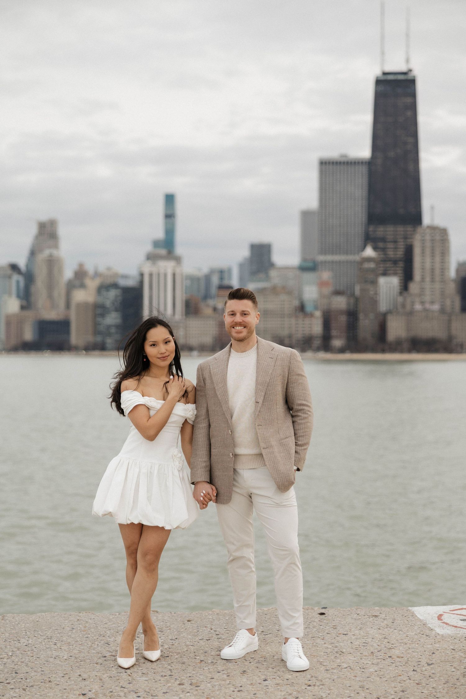 Couple holding hands in white dress and tan suit with Chicago skyline backdrop.