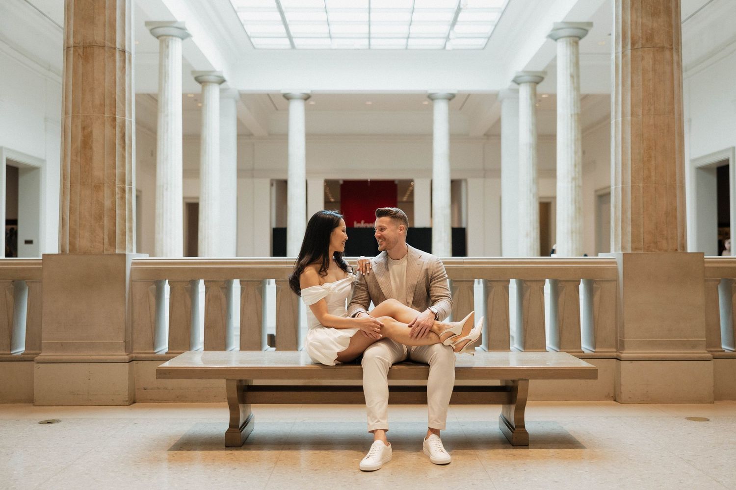 Stylish couple sitting together on a wooden bench in a classical museum atrium with tall columns.
