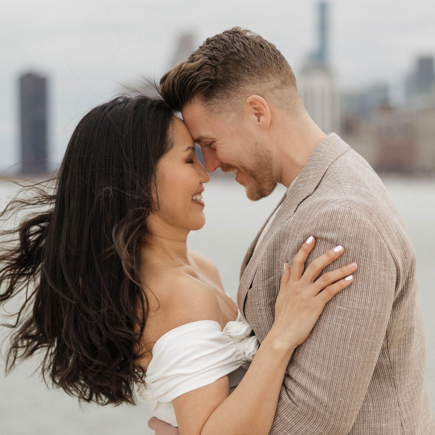 Romantic couple embracing outdoors with city skyline backdrop, woman in white off-shoulder top and man in beige linen shirt.