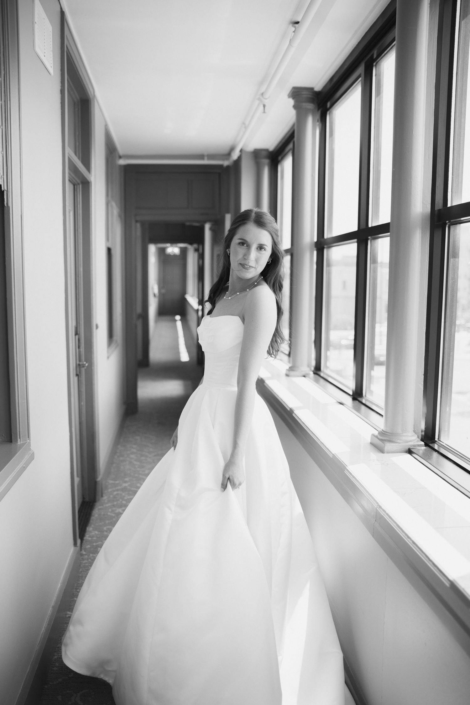 Black and white photo of a bride in a white ball gown standing in a hallway with large windows.