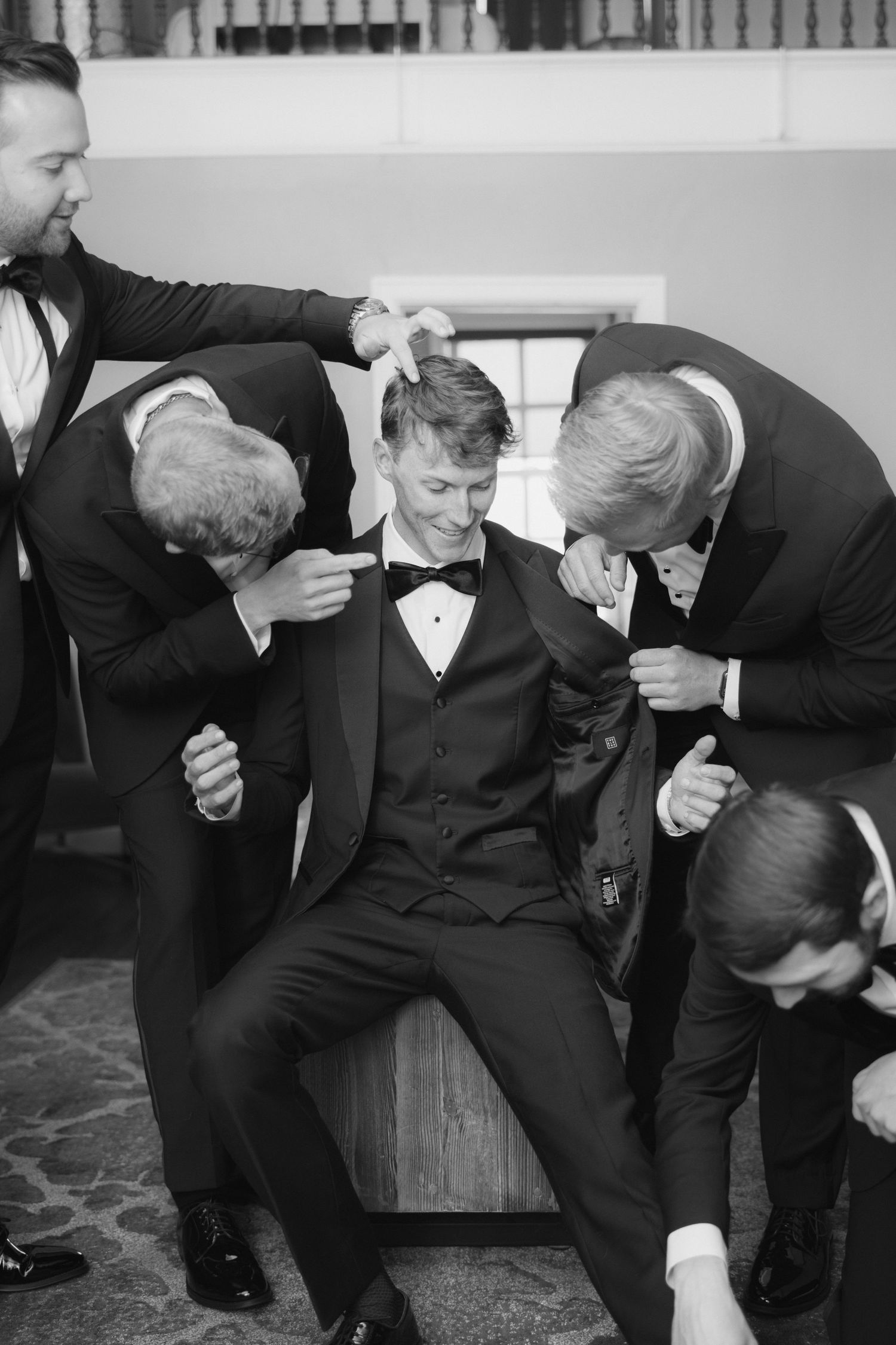 Groomsmen helping groom get ready in tuxedos, black and white wedding photo.