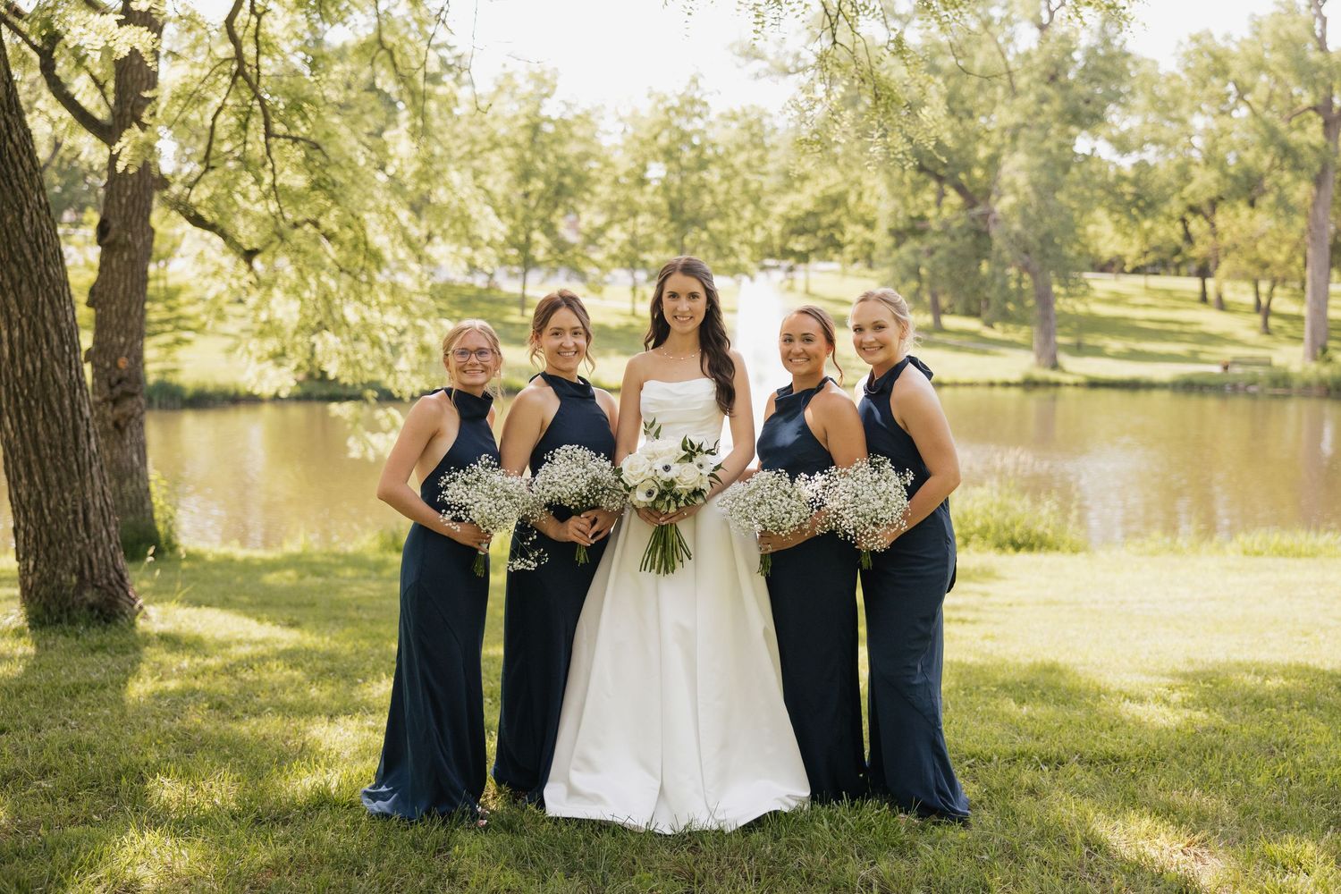 Bride in white gown poses with four bridesmaids in navy blue dresses holding bouquets outdoors near a pond.