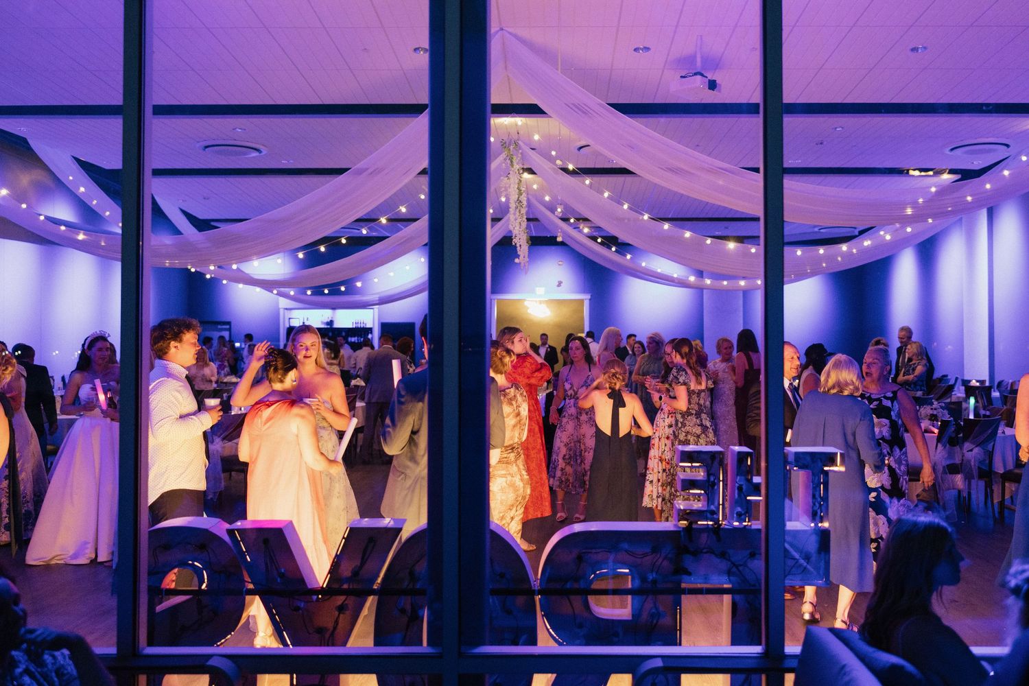Wedding reception viewed through large windows with purple lighting, draped ceilings, and guests dancing on the floor.