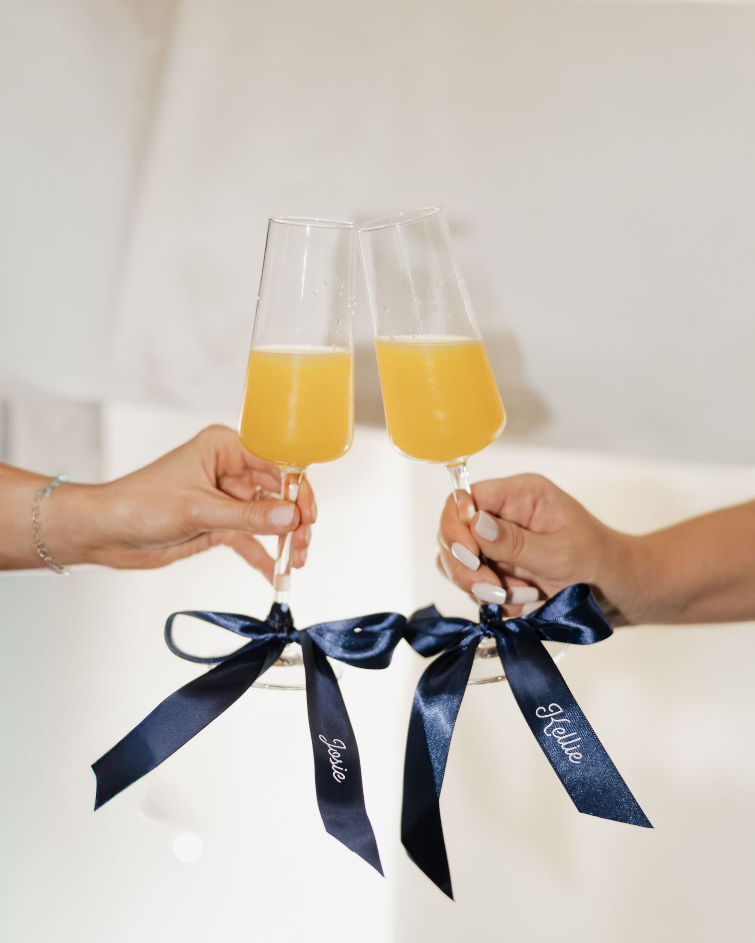 Two champagne flutes filled with mimosas, adorned with navy blue satin ribbons, being toasted by two hands at a wedding.