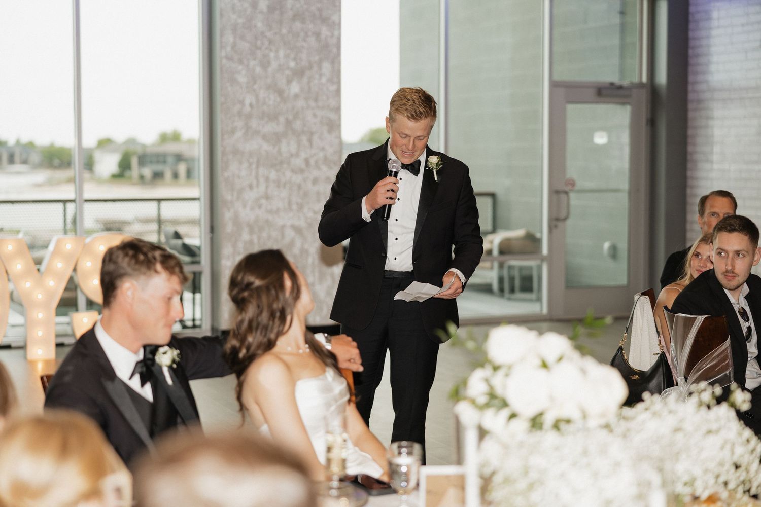 Man in tuxedo giving wedding toast speech at elegant reception while bride and groom listen at decorated head table.