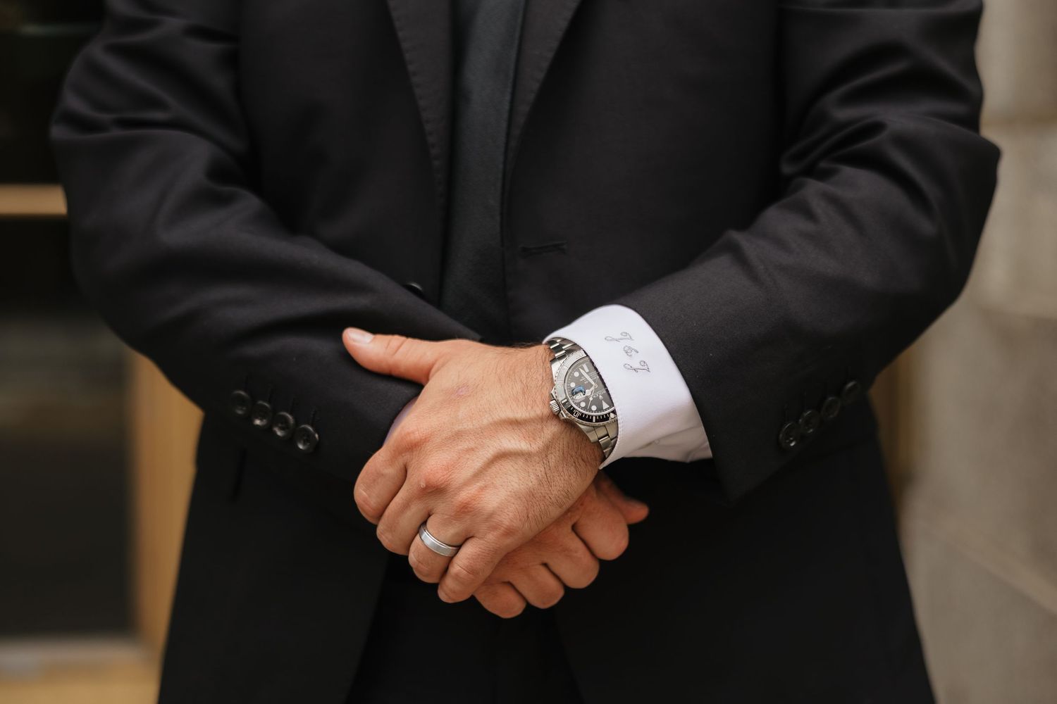 Man in black suit with clasped hands wearing a large silver skeleton watch and gold ring.