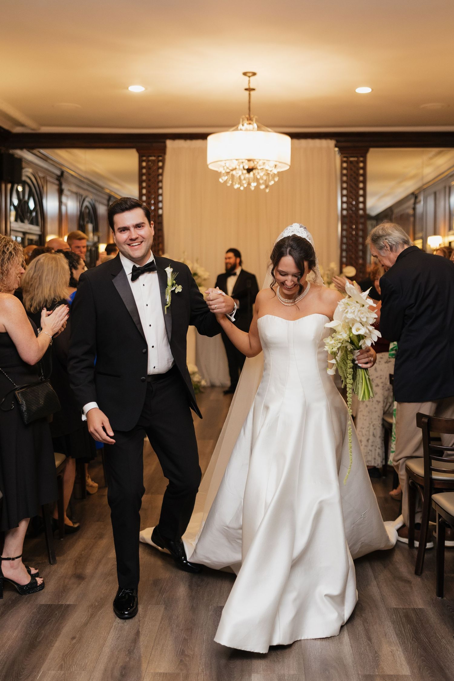 Bride and groom walk joyfully through reception hall, she holds white bouquet, he wears black tuxedo, guests applaud around them.